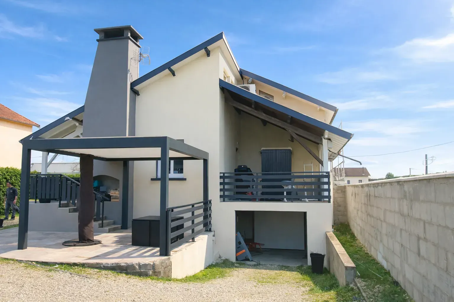 Une maison claire de deux étages avec une cheminée grise, une terrasse extérieure, un balcon et un garage de plain-pied, sous un ciel bleu.