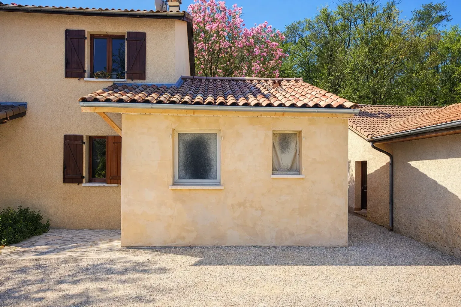 Une extension de maison de plain-pied, de couleur beige, avec un toit de tuiles en terre cuite, située sur une allée de gravier sous un ciel d'un bleu éclatant.