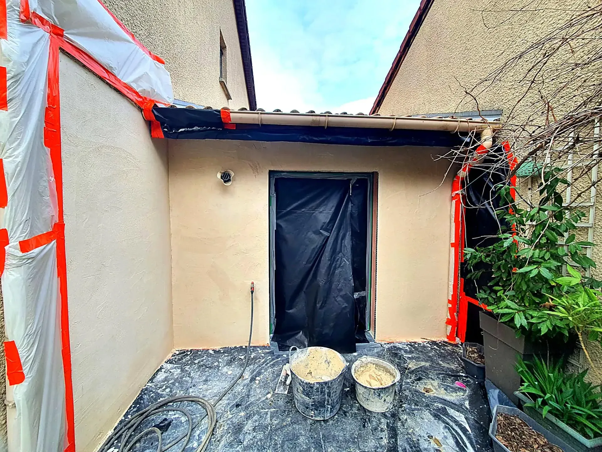 Sur un chantier, on aperçoit une porte recouverte de plastique noir, deux seaux sur une bâche et des murs masqués par du ruban adhésif rouge.
