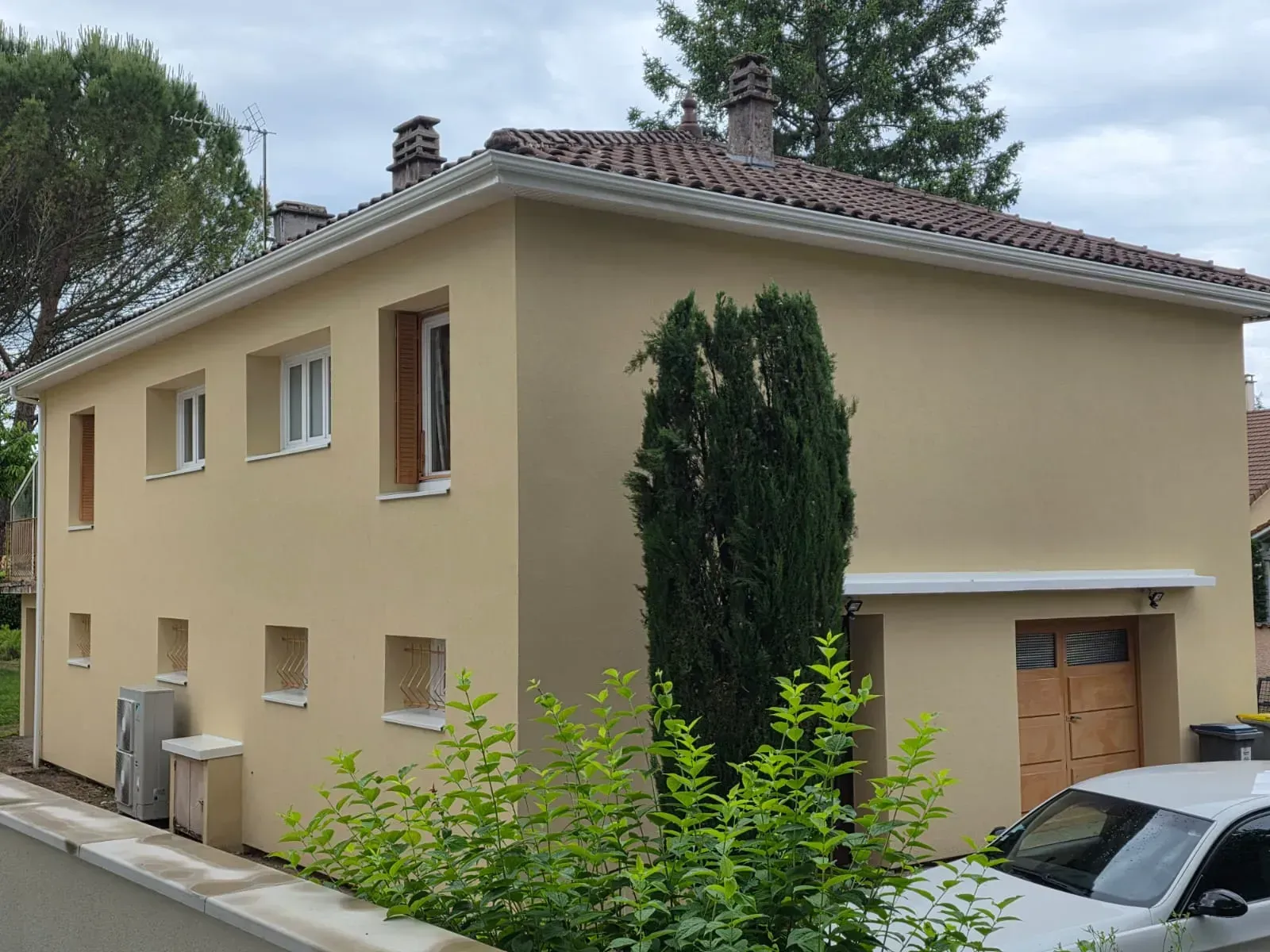 Une maison beige à deux étages avec un toit de tuiles, de petites fenêtres et un garage, vue de la rue près d'un arbre vert.