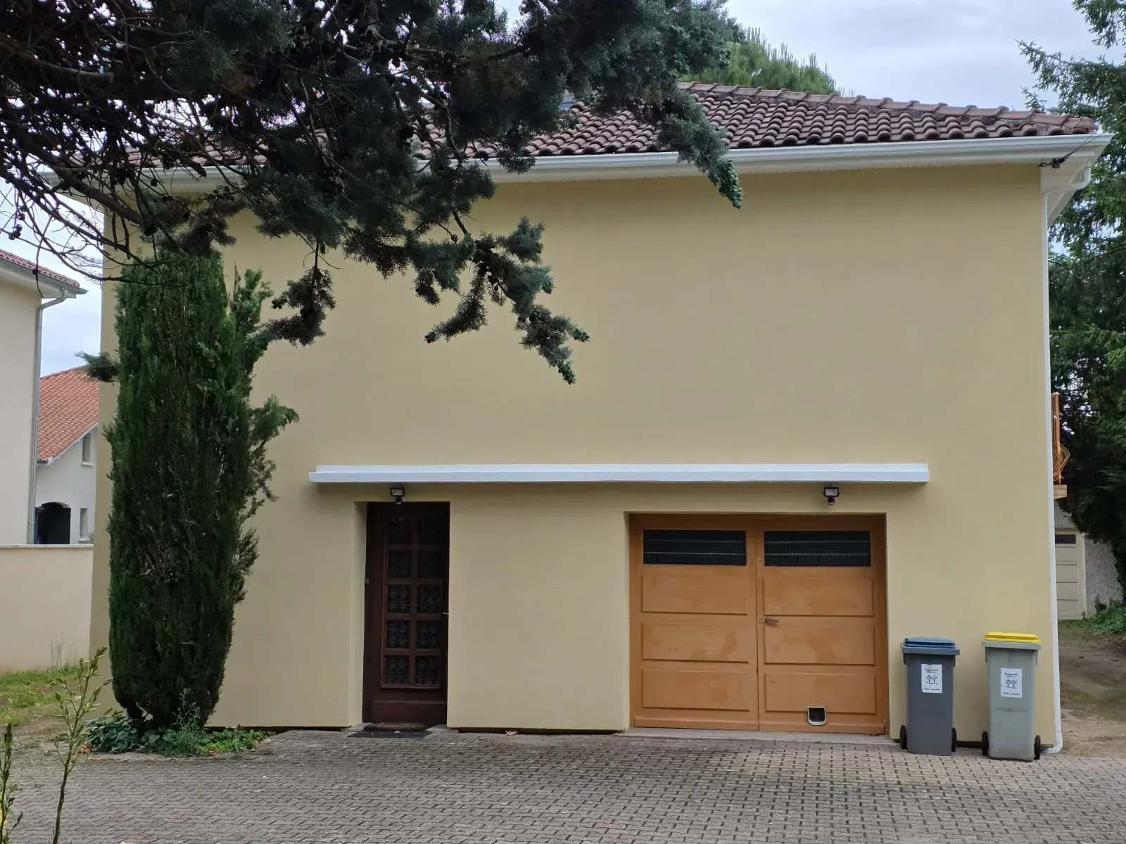 Une maison jaune pâle à deux étages avec une porte de garage en bois, une porte latérale et deux poubelles sur une allée pavée.