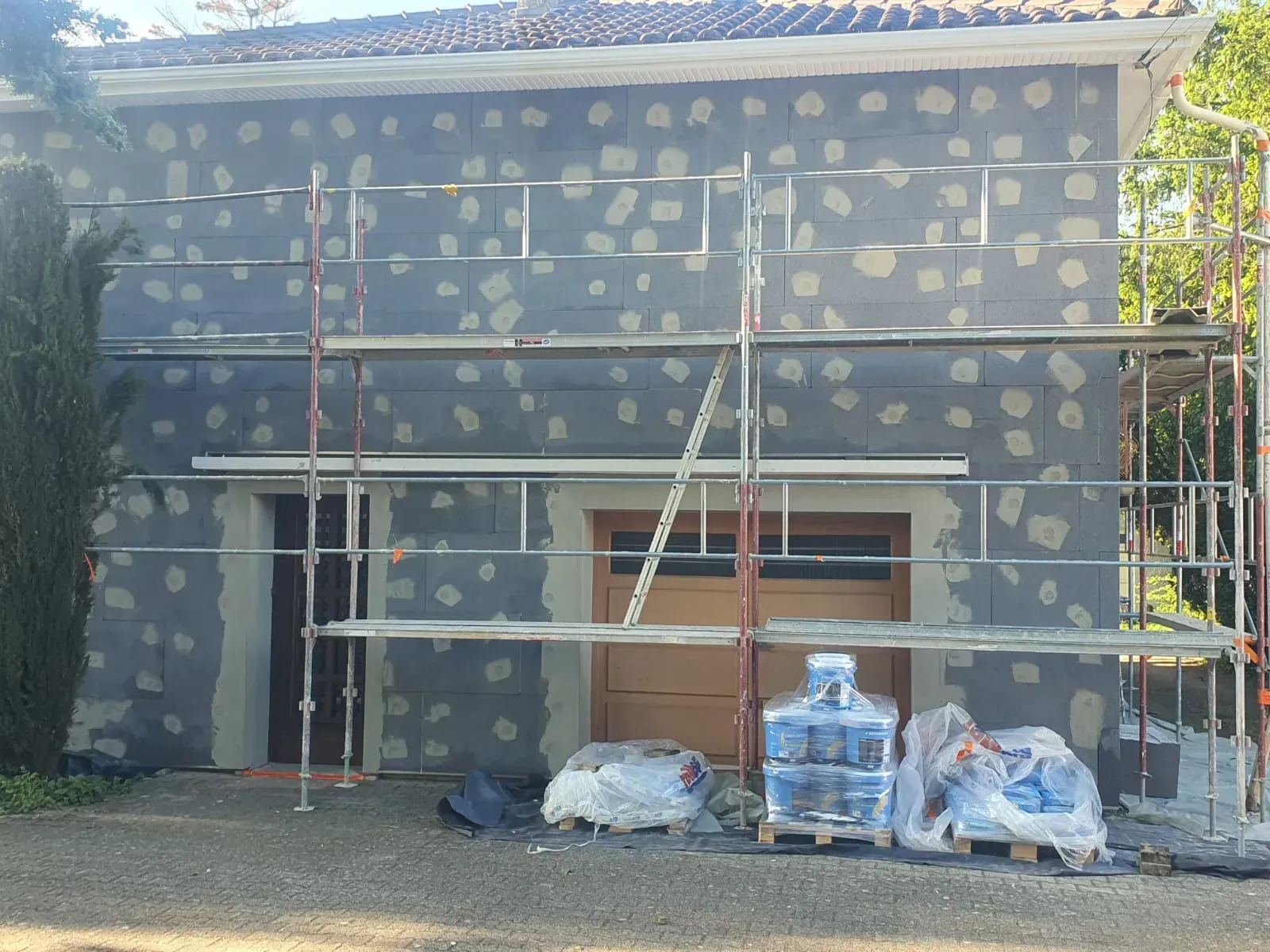 Des échafaudages se dressent devant un bâtiment dont le mur gris foncé présente des plaques de plâtre claires régulièrement espacées.