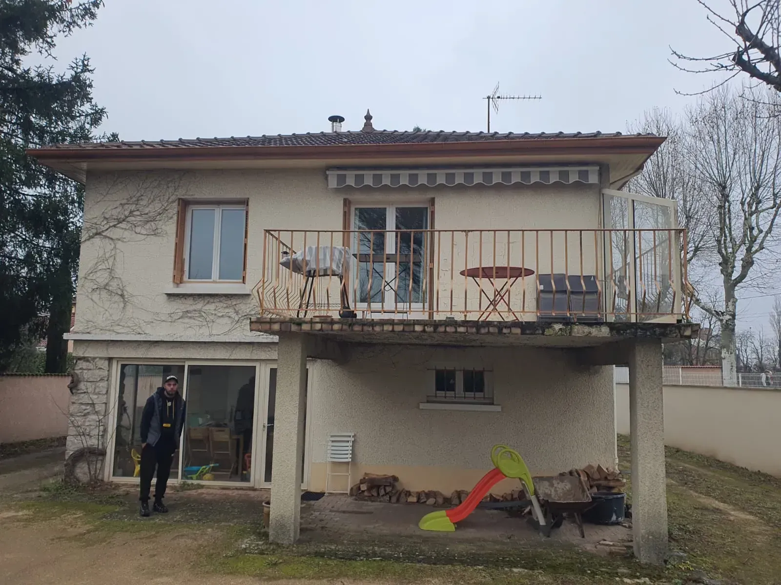 Une personne se tient devant une maison beige à deux étages avec un balcon surélevé et une terrasse agrémentée d'un petit toboggan pour enfants.