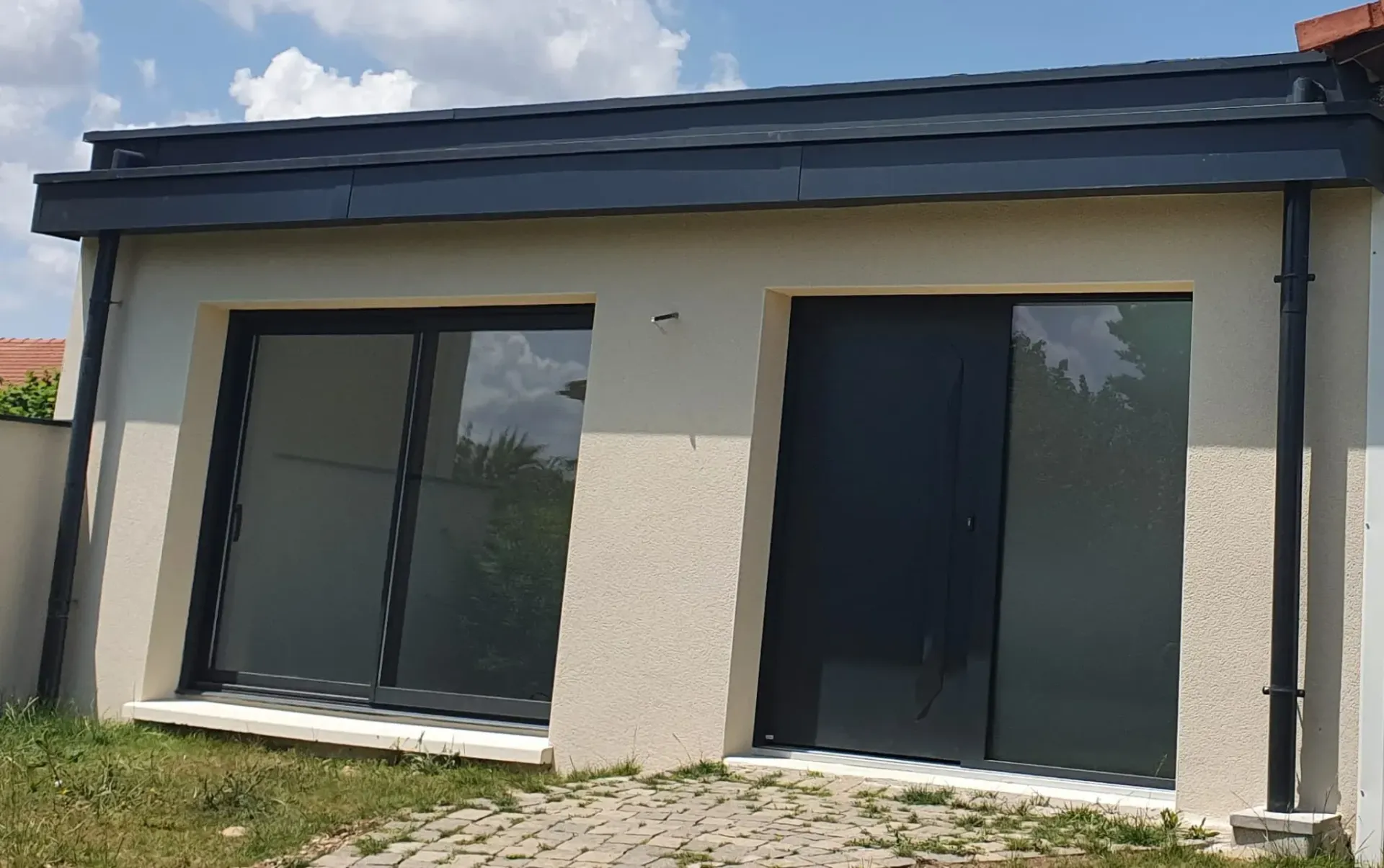 L'extérieur d'un bâtiment moderne et clair, avec un toit foncé, des gouttières noires, une grande porte coulissante en verre et une porte pleine.