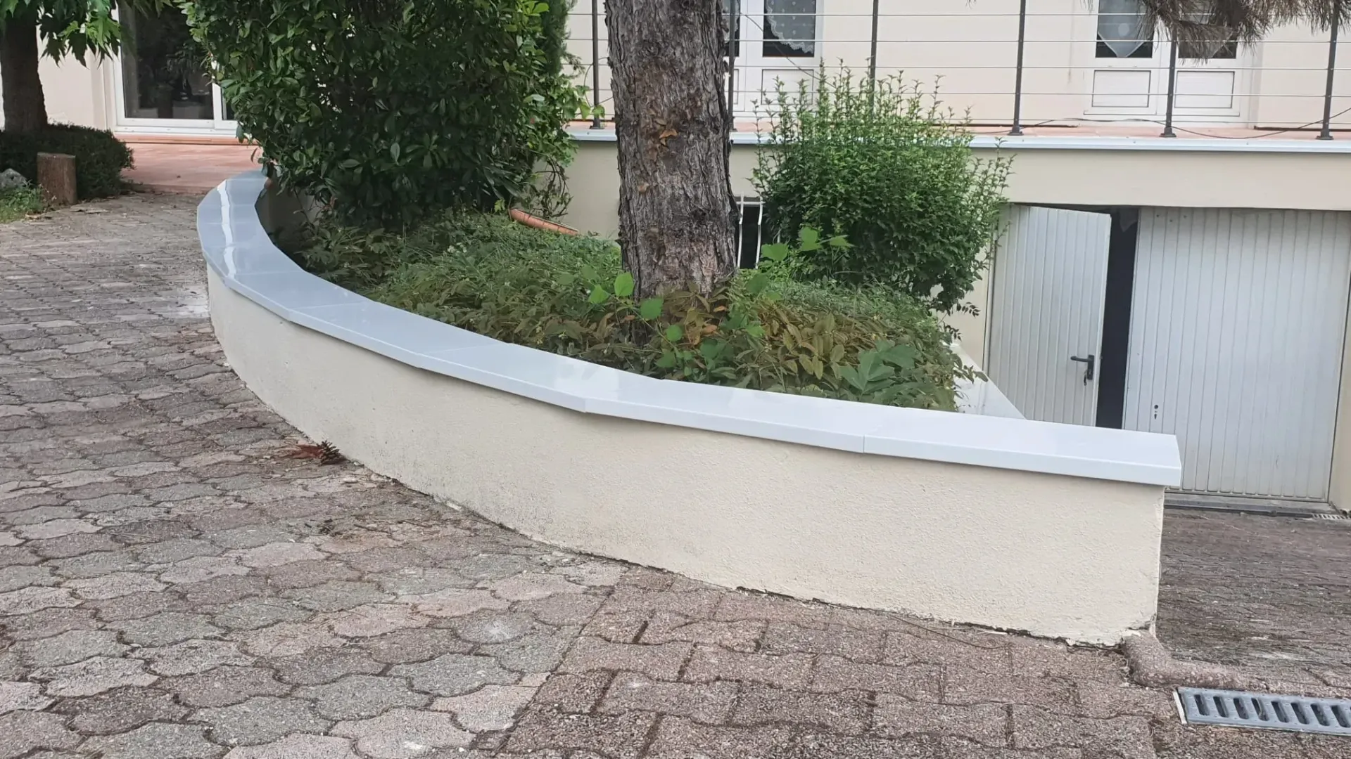 Un muret de jardin beige incurvé, coiffé d'un chapeau blanc, entouré d'une allée pavée, d'arbustes verts et d'un arbre.