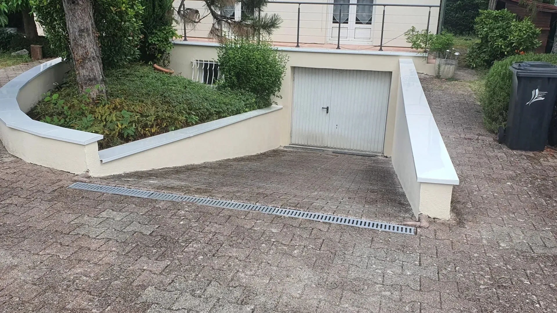 Une allée en béton descend en pente douce vers un garage souterrain, à côté d'un muret de soutènement agrémenté d'aménagements paysagers et d'un grand arbre.