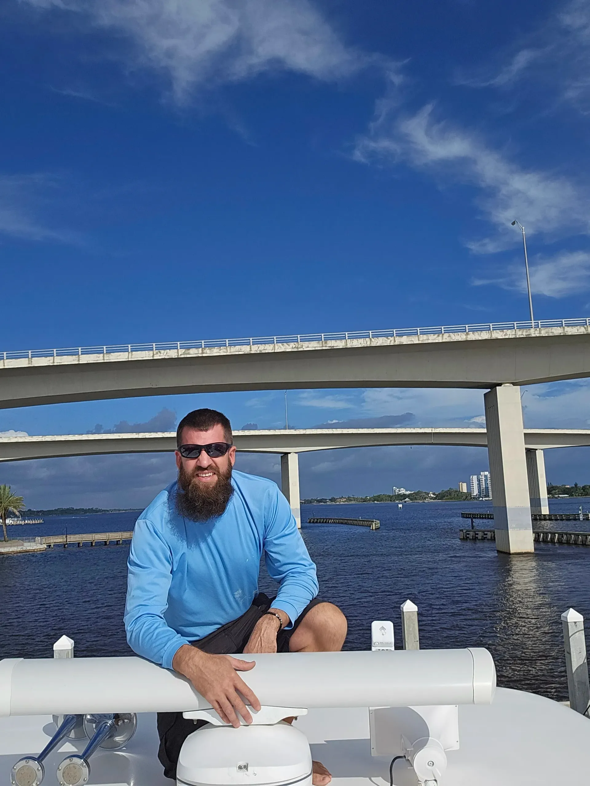 Man on a boat wearing sunglasses, blue shirt, near a bridge over water on a sunny day.