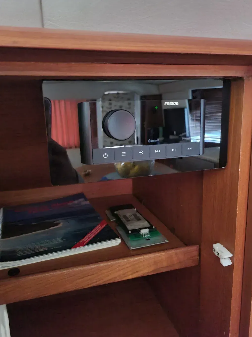 A radio mounted in a wooden cabinet. Below it, a shelf with a book and circuit board.