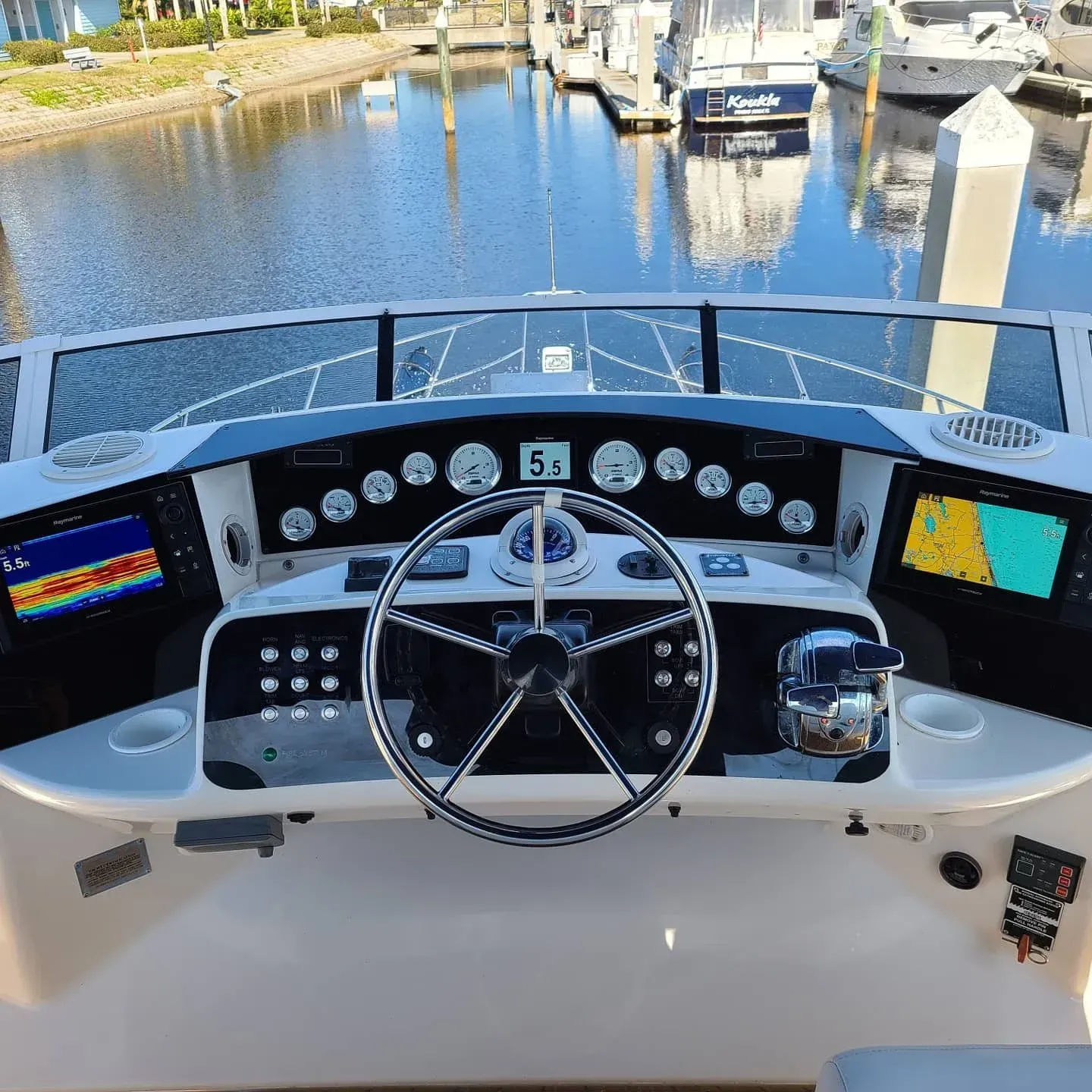 Boat's helm: steering wheel, instruments, displays, and a view of the marina in sunny conditions.