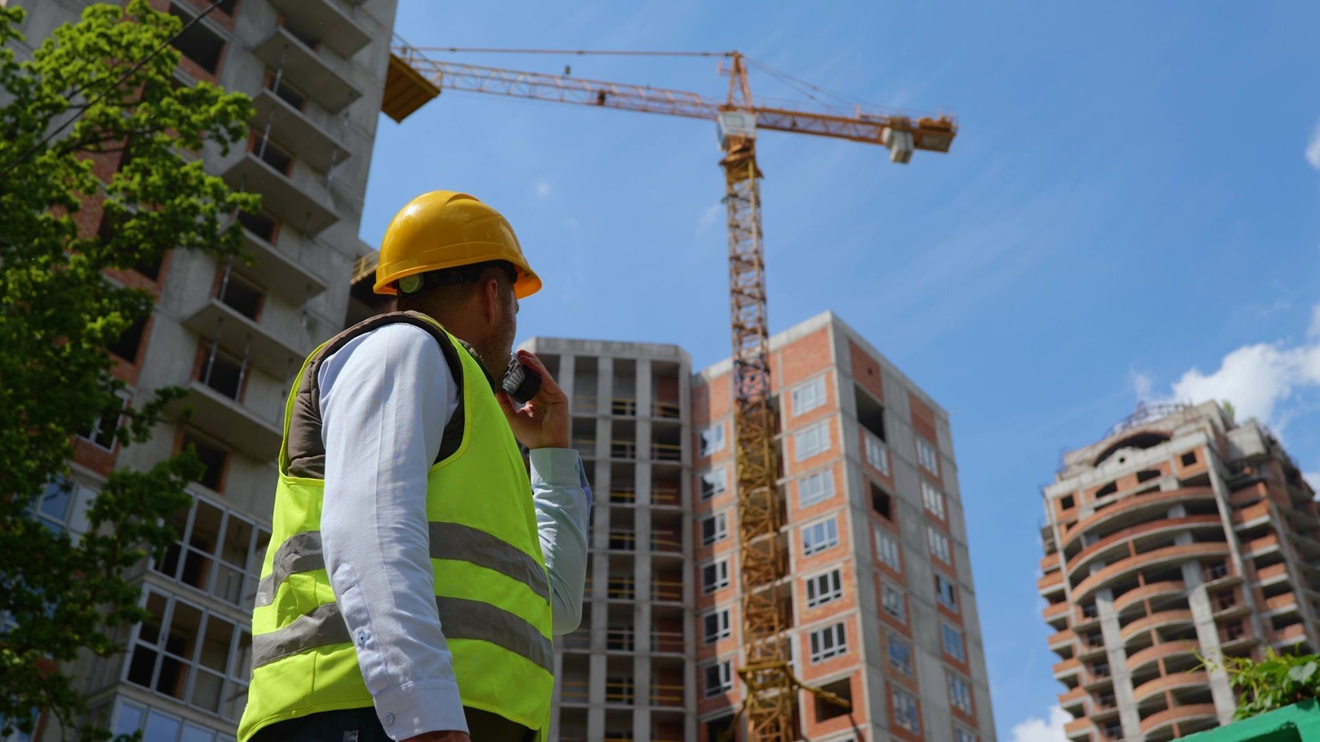 Low-angle view of a builder in a helmet using a walkie-talkie, standing at a city construction site.