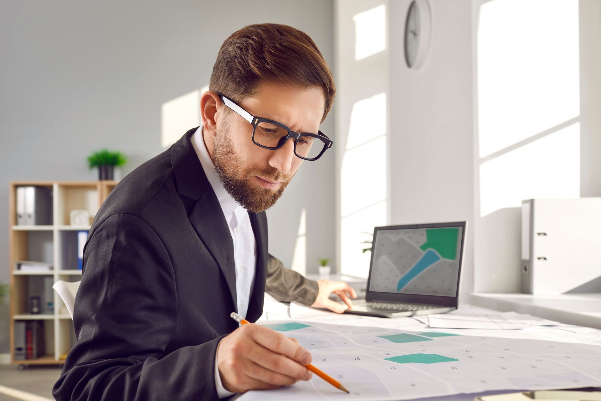 A surveyor sitting at an office table working with cadastral maps and plans on a laptop.