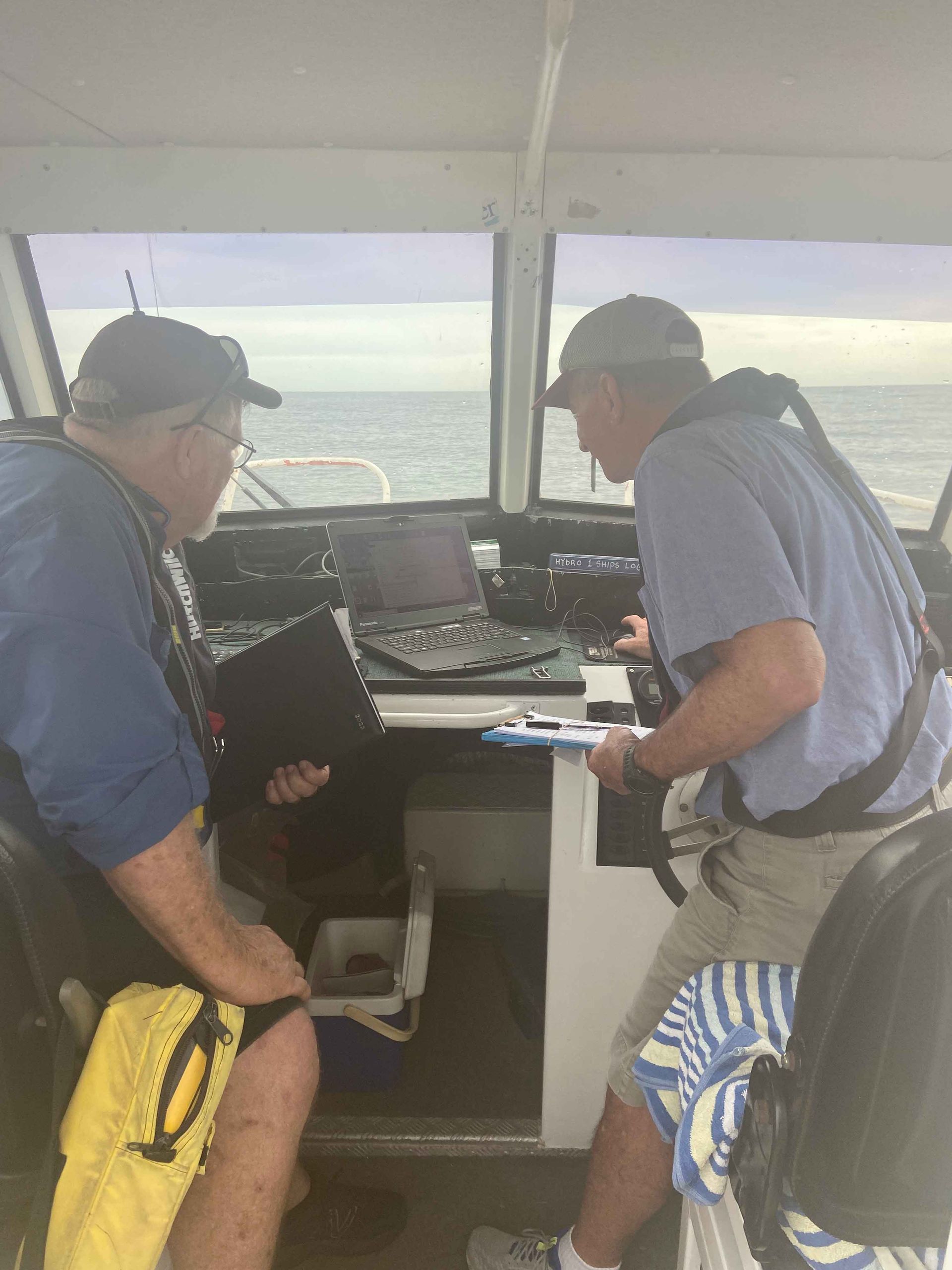 Two men in life vests inside a boat's cabin, looking at a screen, with ocean visible through windows.