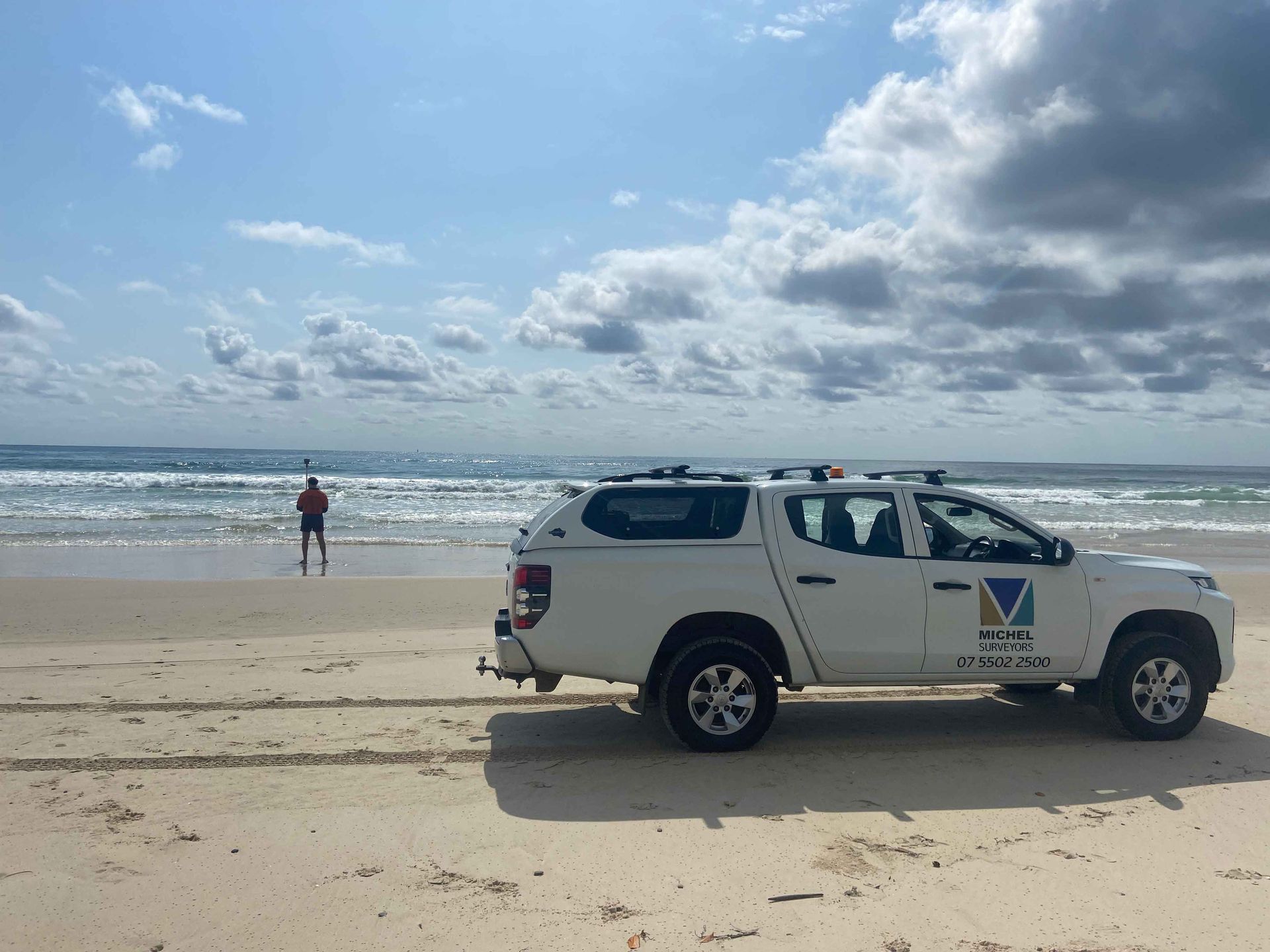 White truck on beach, person standing in water, blue sky, and waves.