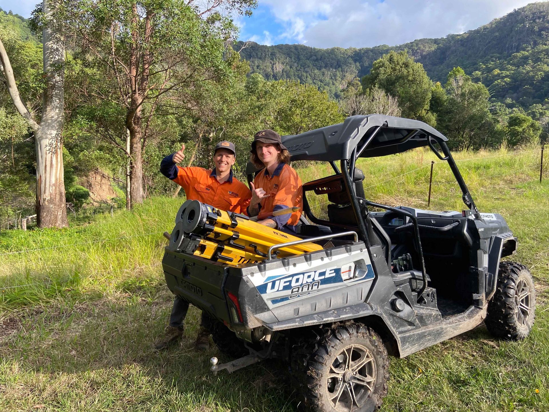Two people in orange shirts give thumbs up, standing by an off-road vehicle in a grassy field, mountains in the background.
