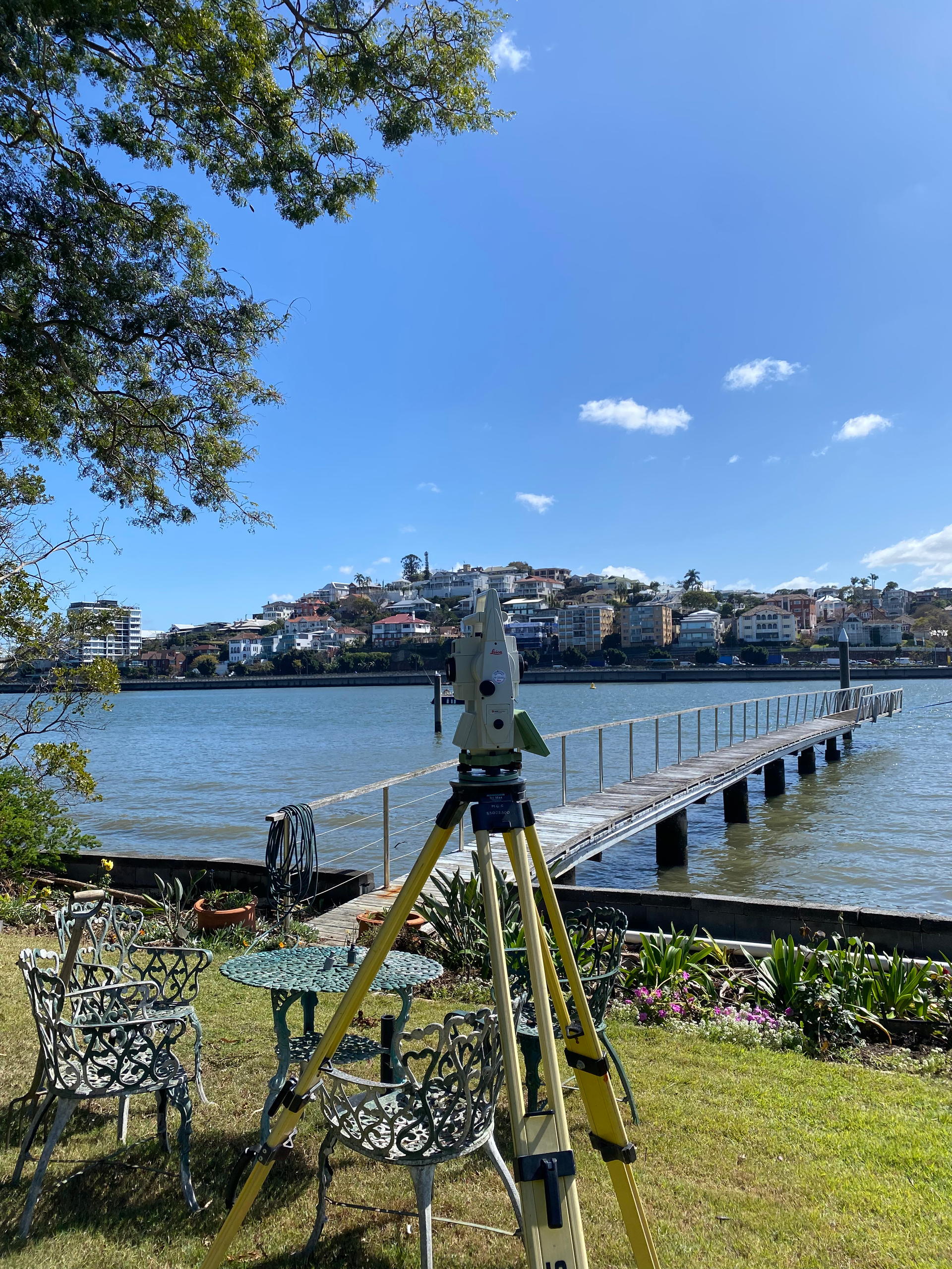Surveying instrument on a tripod by a waterfront, looking towards a pier and buildings on a sunny day.