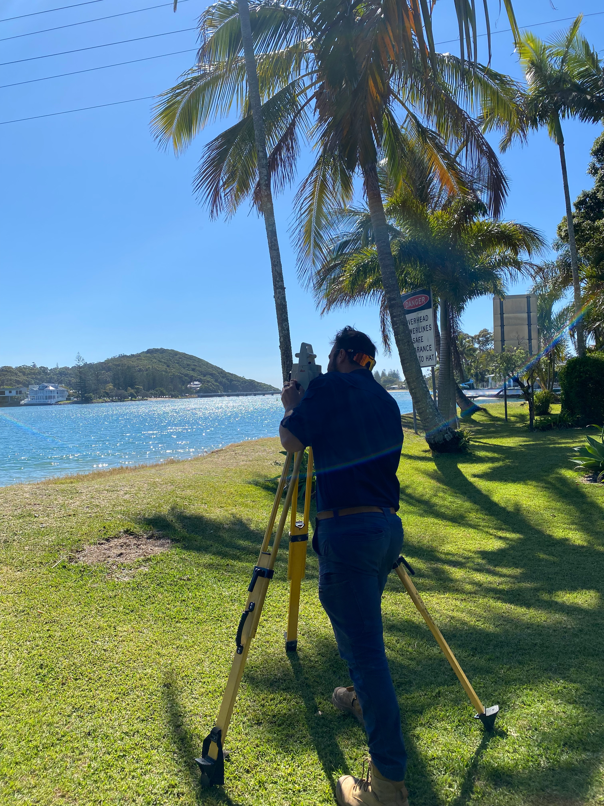 Man surveying by the water, using a tripod and measuring device. Palm trees, blue sky, and a boat in the distance.