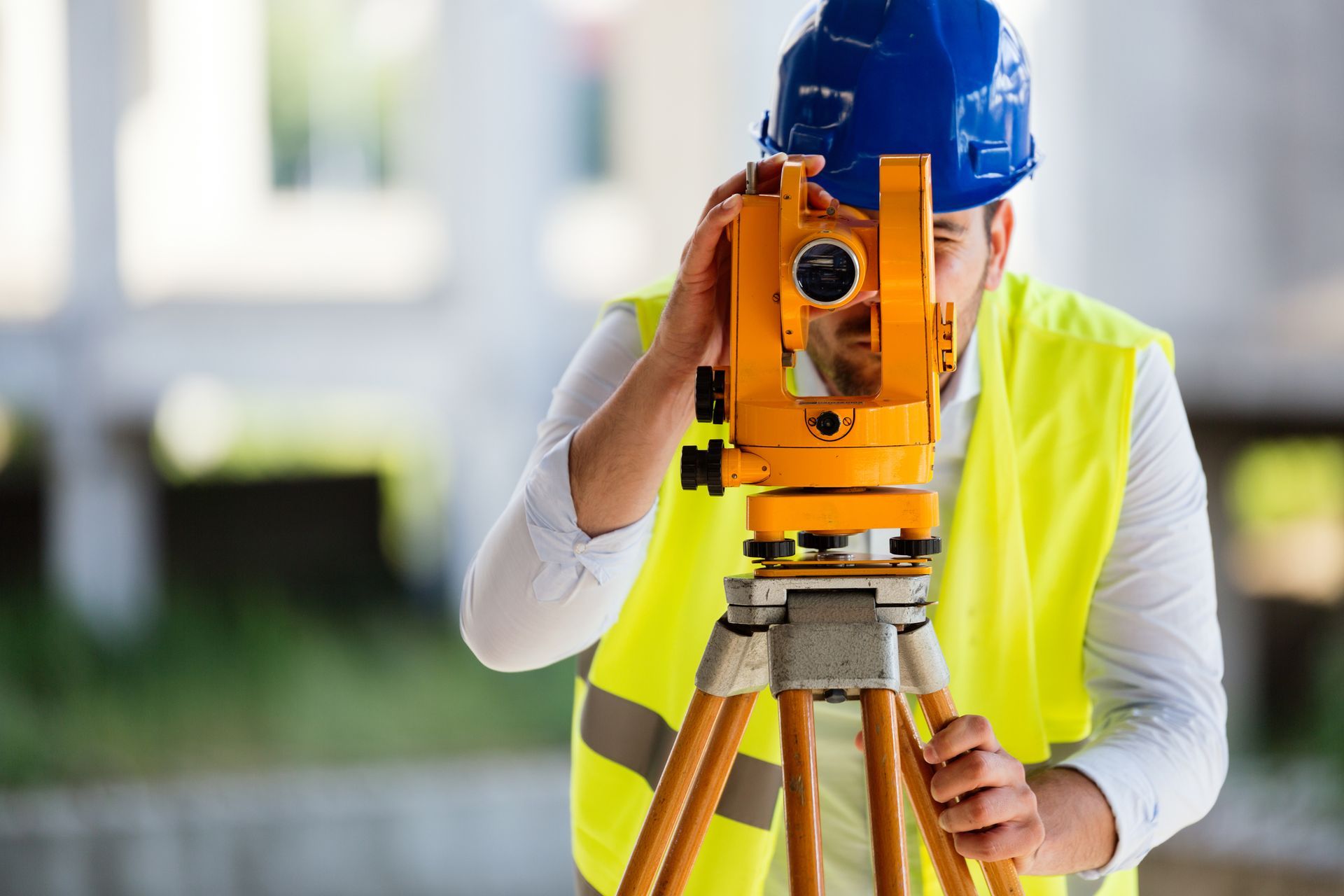 A land surveyor in a safety vest and blue helmet, working on a building site and using equipment.