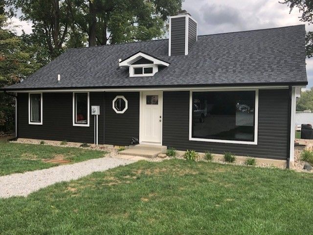 Dark-sided house with white trim, chimney, and door; grey gravel path. Cloudy sky, green lawn.