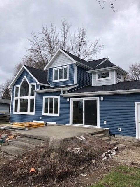 Blue-sided house with a dark roof and large windows under a cloudy sky. A concrete patio is in front.