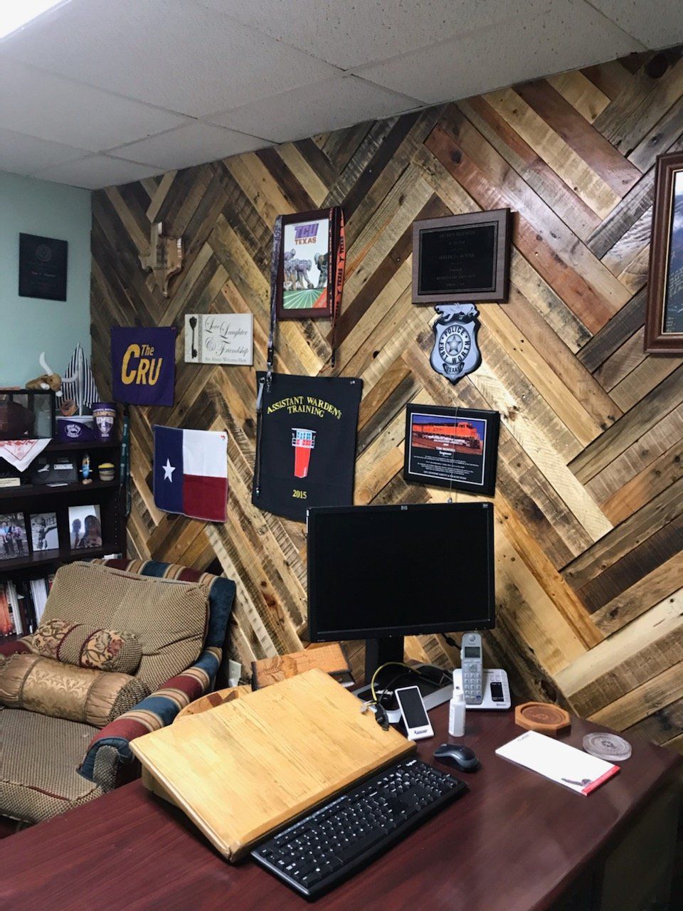 Office with wood-paneled wall, desk, computer, diplomas, flags, and a comfortable chair.