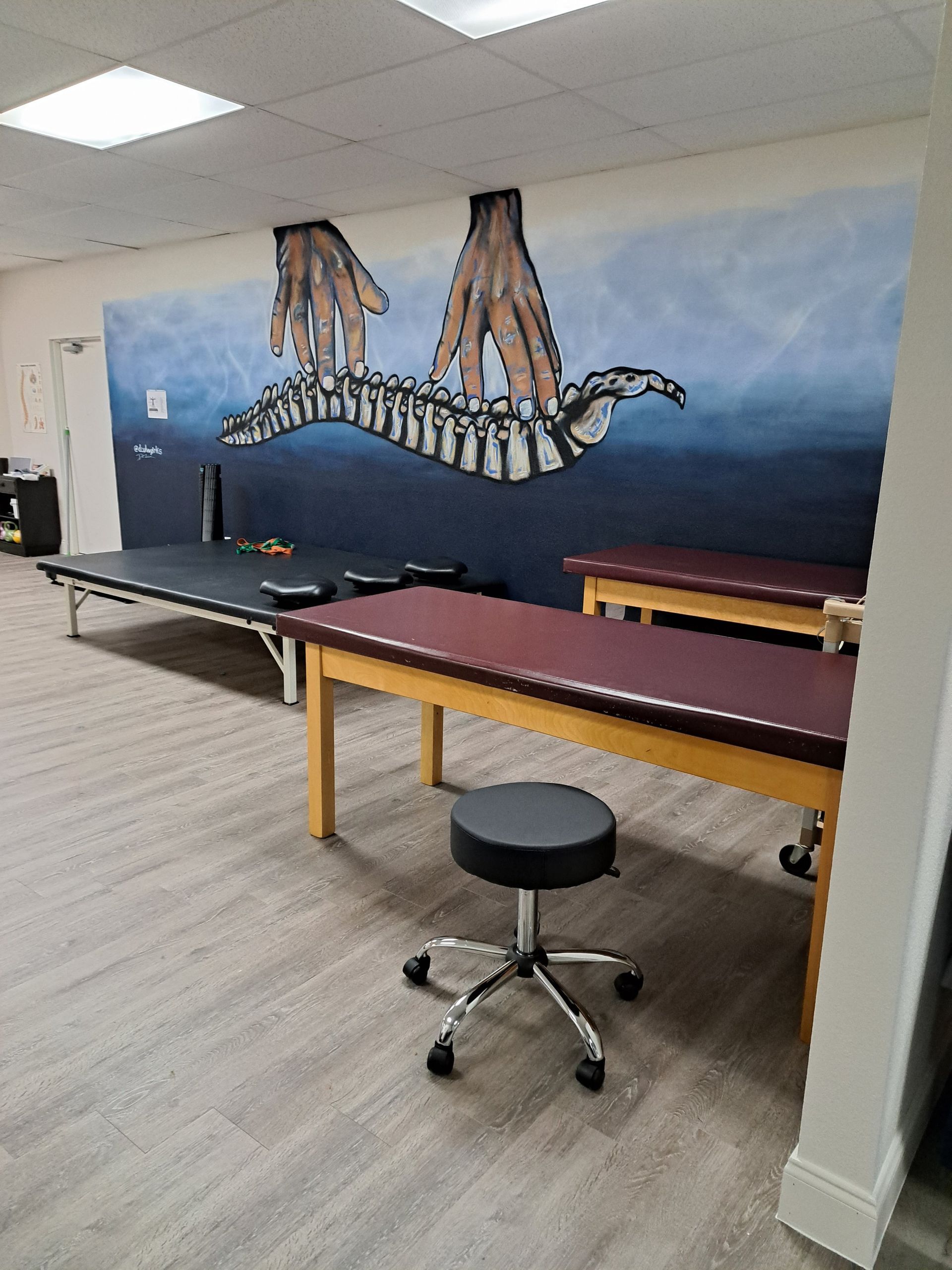 Physical therapy room with treatment tables, a stool, and a spine-themed mural on the wall.