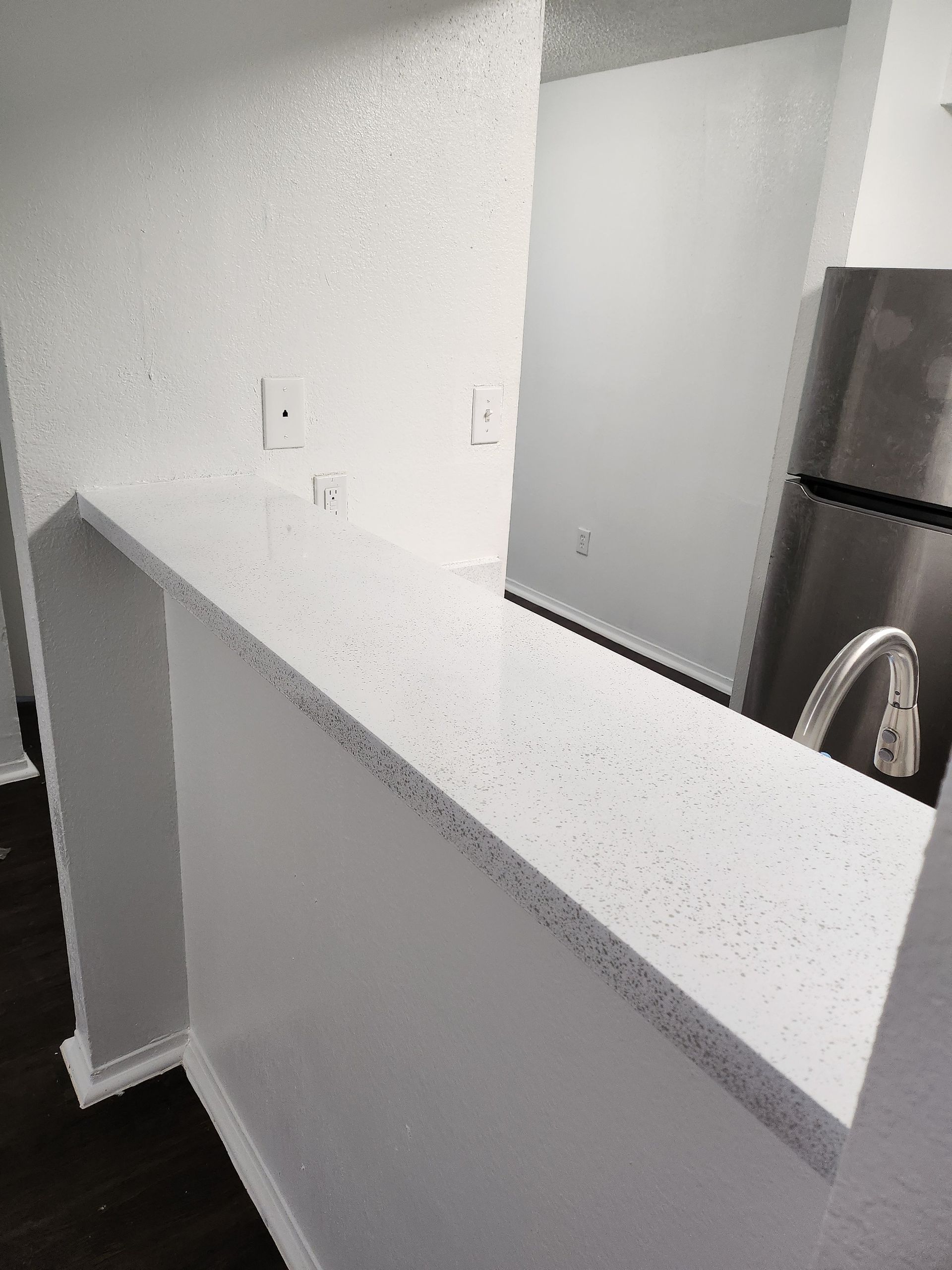 White countertop with flecks and wall in a kitchen, stainless steel refrigerator visible.