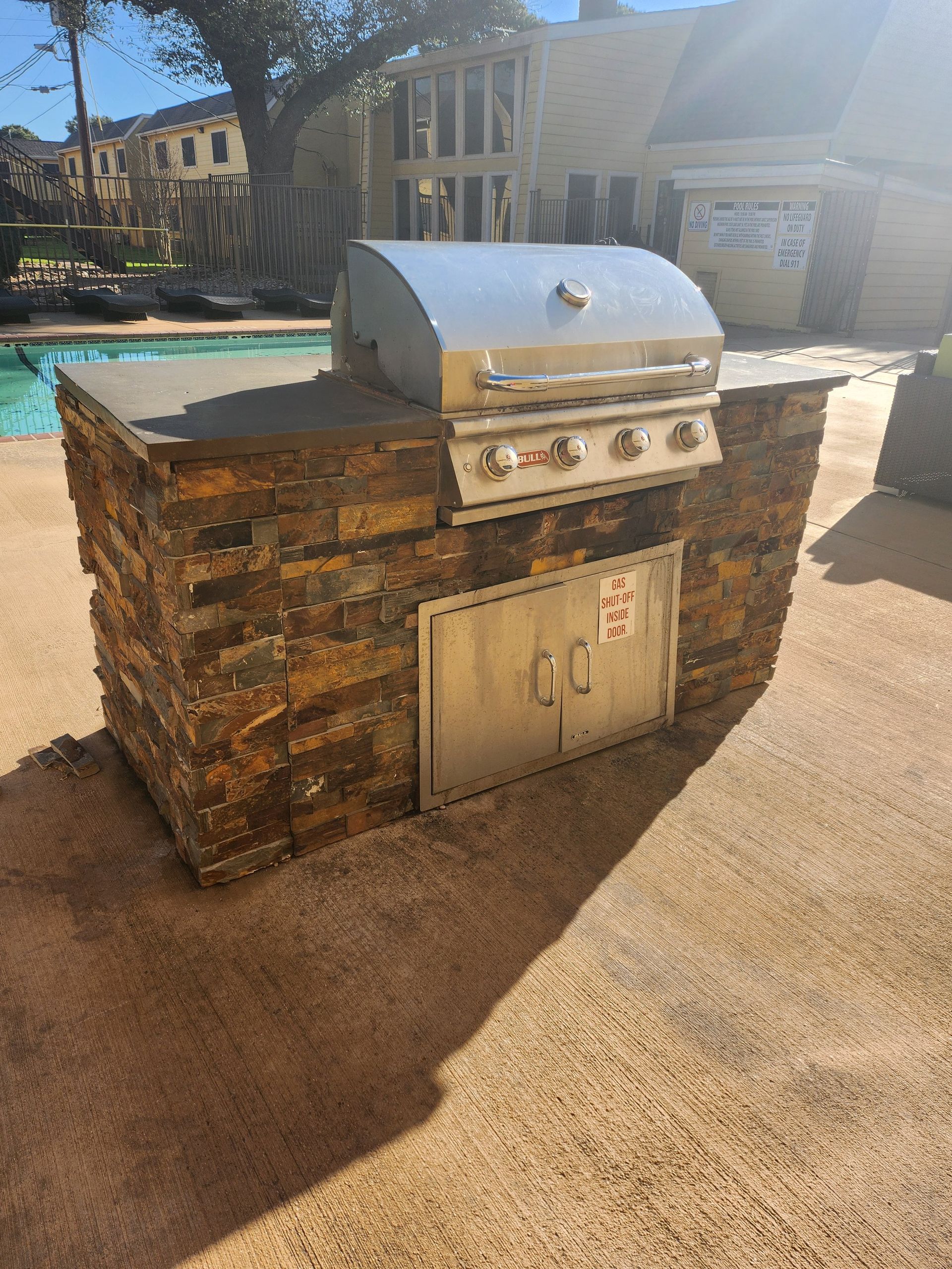 Outdoor built-in stone grill with a stainless steel grill top and storage doors near a pool.