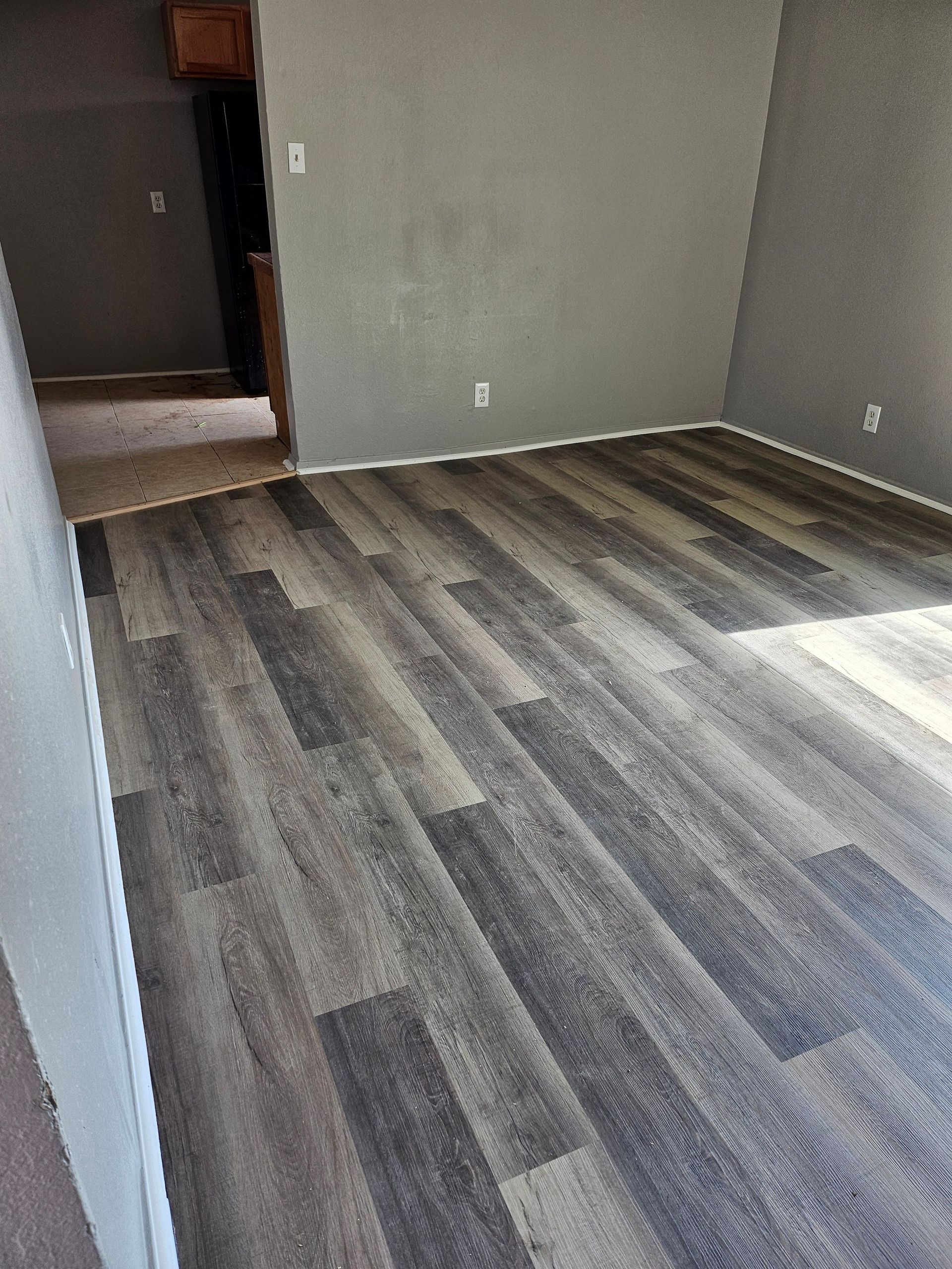 Gray-painted room with new gray wood-look flooring. Entryway to a dark room visible in the background.