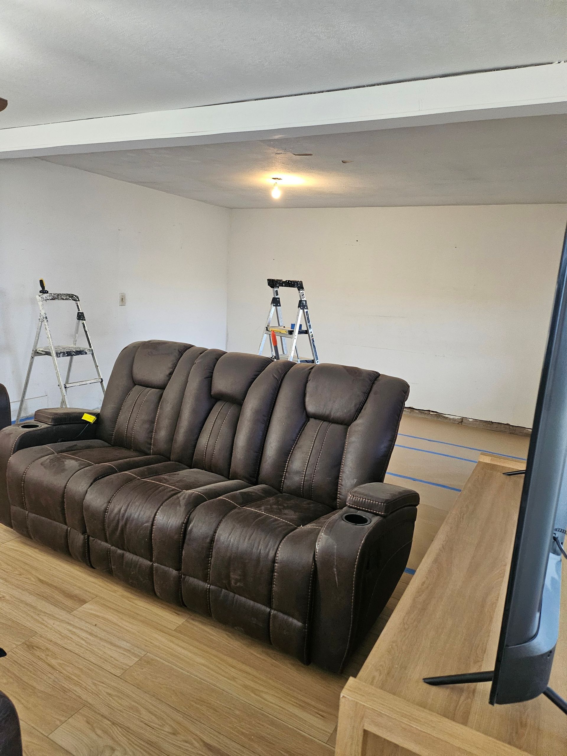 Brown leather reclining sofa in a room with light-colored walls and wood flooring. A television and ladders are visible.