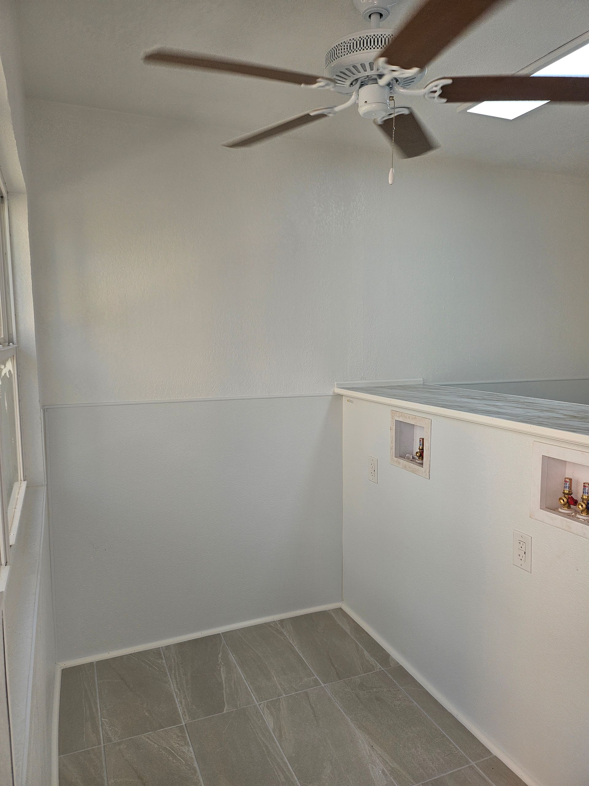 White-walled laundry room with a ceiling fan, a window, and gray carpet.  Plumbing connections visible.