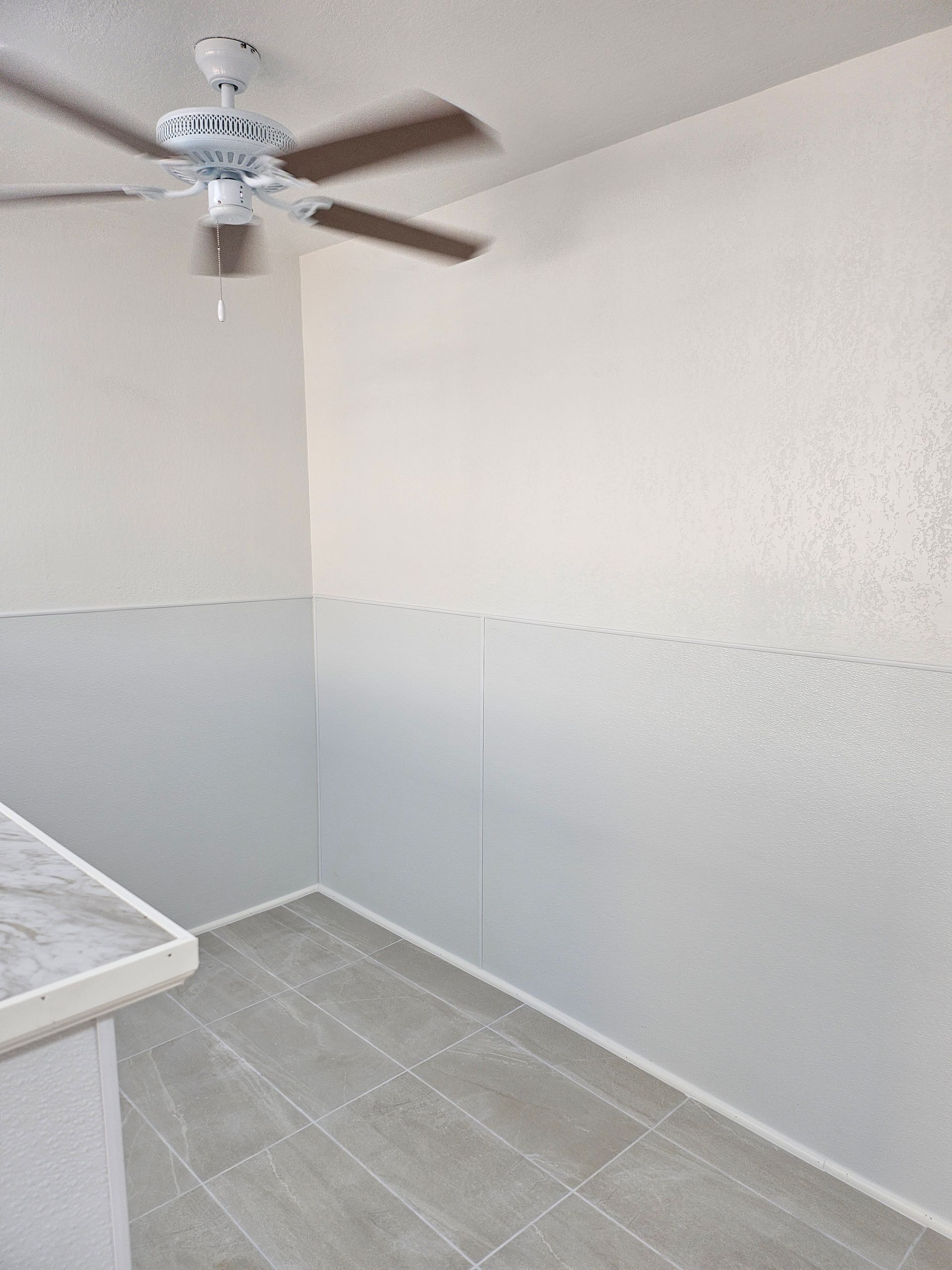 Corner of a room with white walls, ceiling fan, and gray tiled floor.