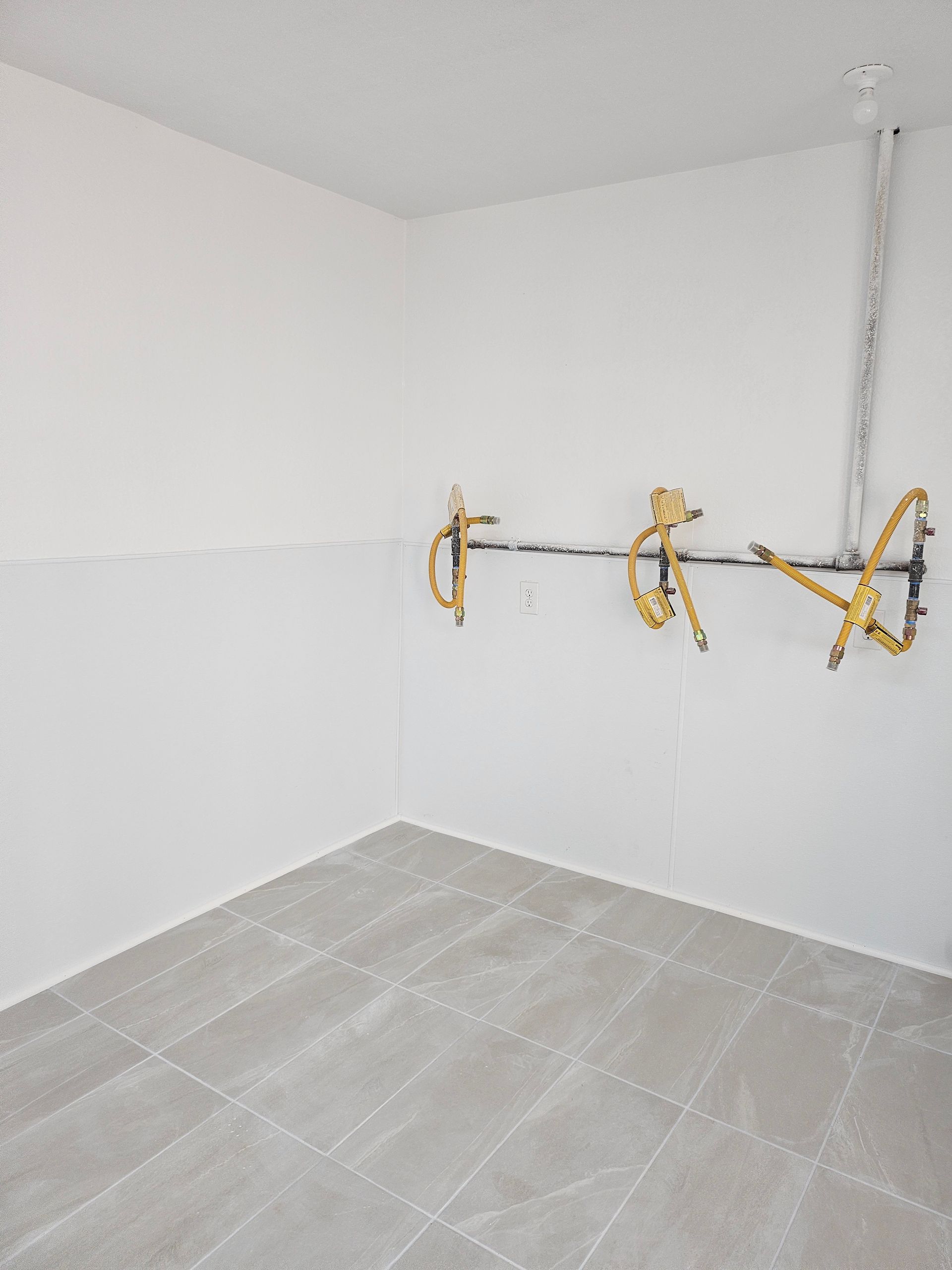 Empty room with exposed plumbing on a white wall, gray tiled floor.