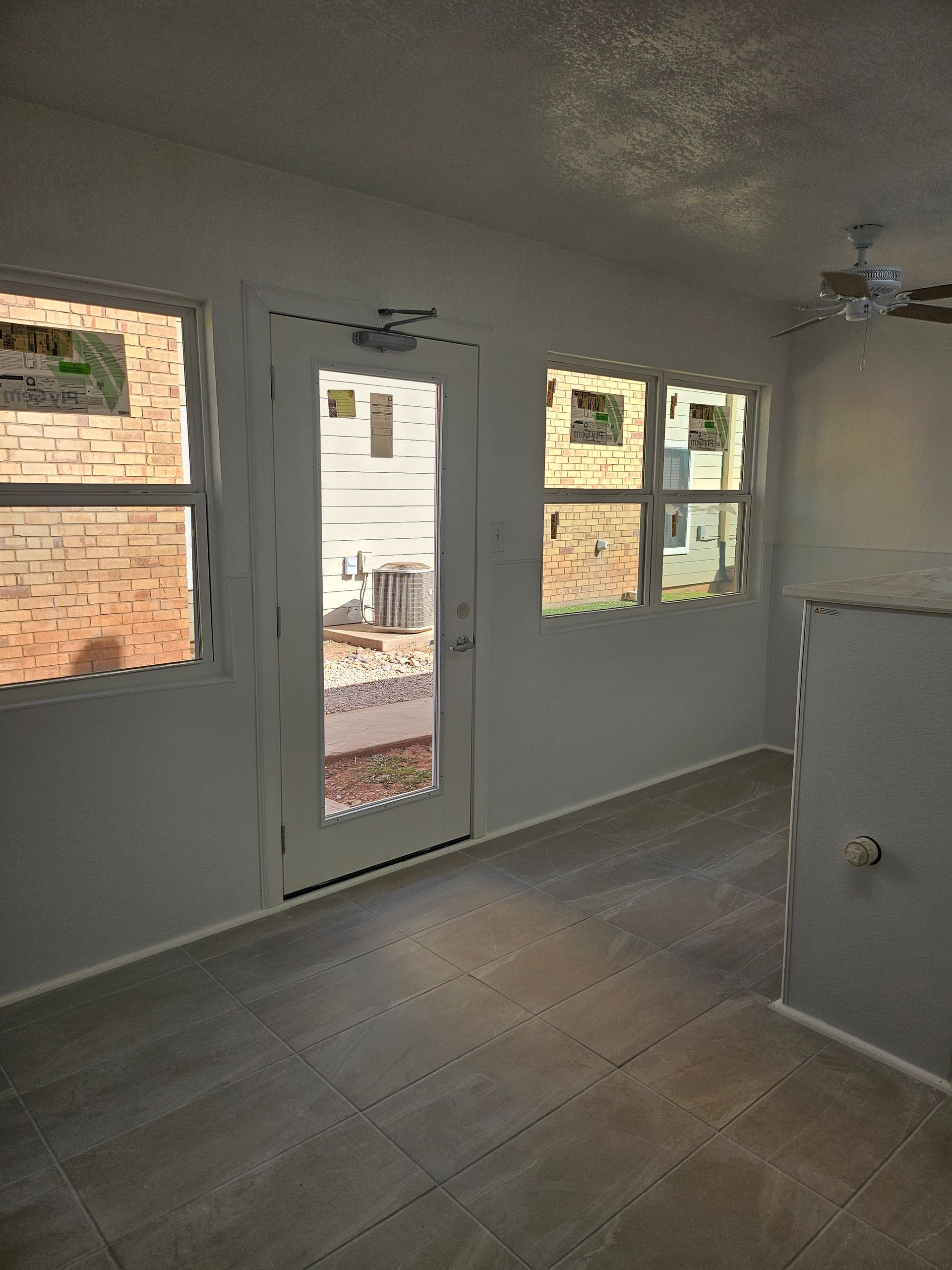 Interior room with windows, door, and ceiling fan. Light-colored walls and flooring; outside views visible.