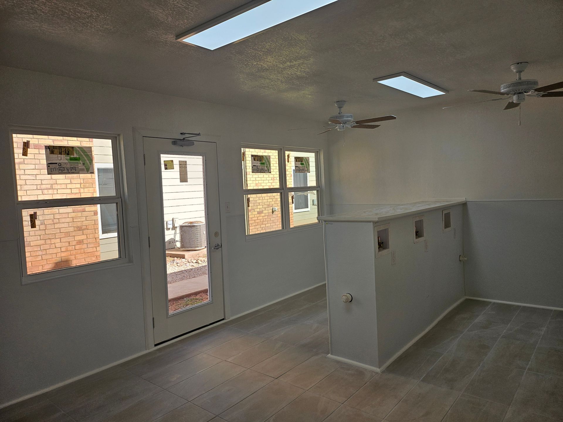 Interior view of a room with windows, a door, and a low wall. Ceiling lights and fans are visible.