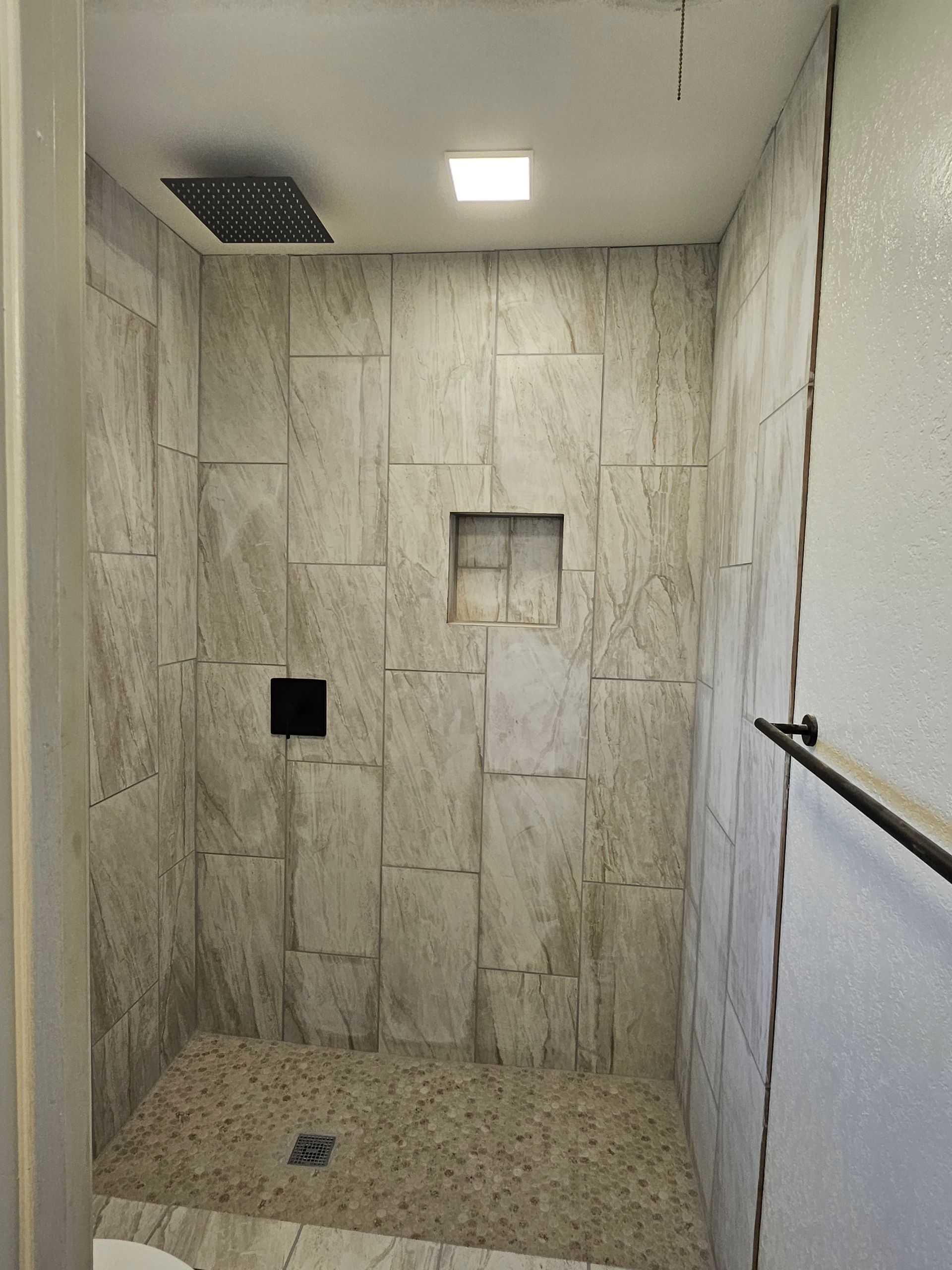 A small, tiled shower with a pebble floor, showerhead, and a shelf built into the wall.