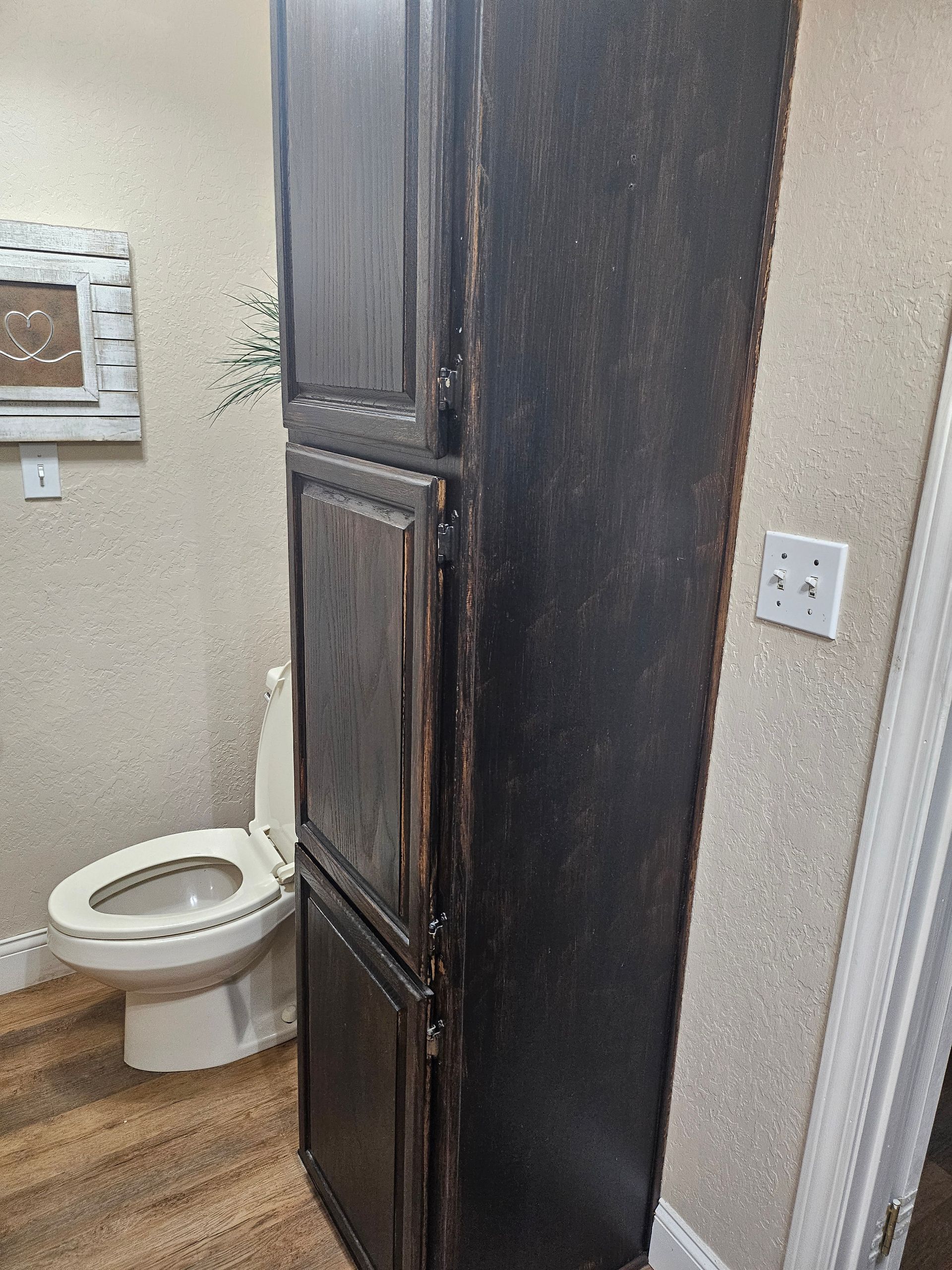 Tall, dark-stained storage cabinet next to a toilet in a bathroom with textured wallpaper and wood-look flooring.