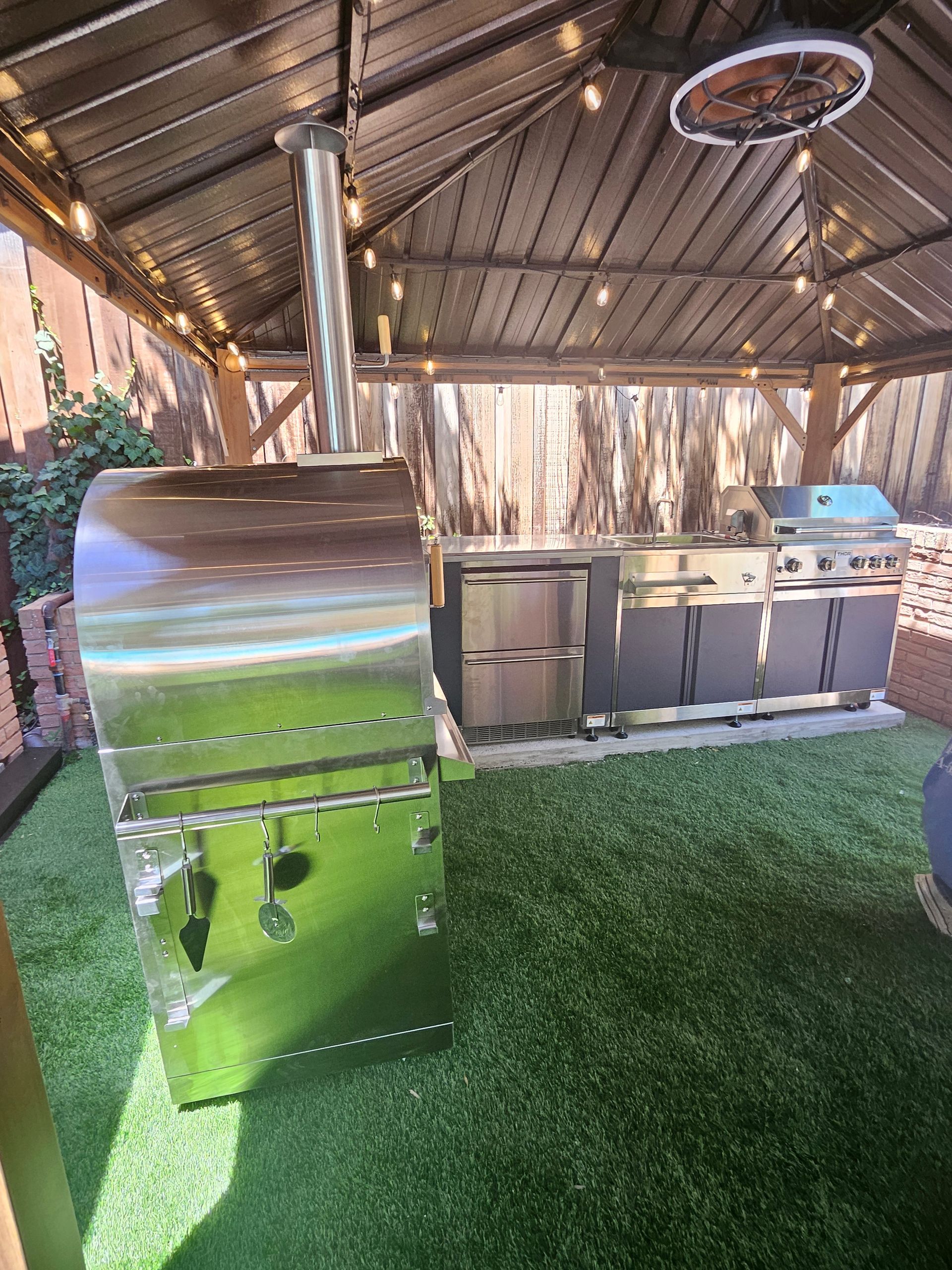Outdoor kitchen with stainless steel grill, pizza oven, and seating under a gazebo. Artificial turf.