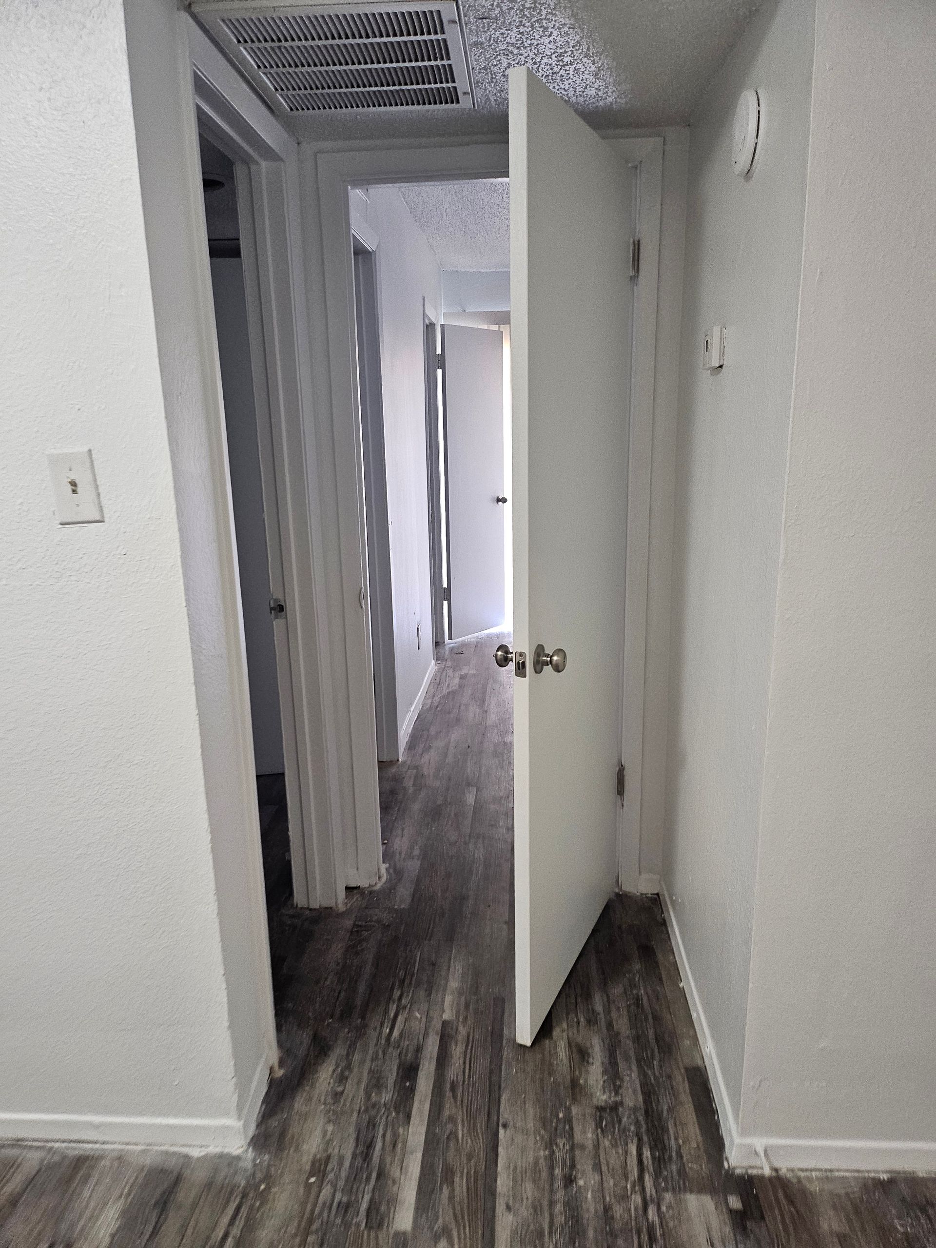 Hallway with wood-look flooring, white doors, and walls. One door is partially open, leading to another.