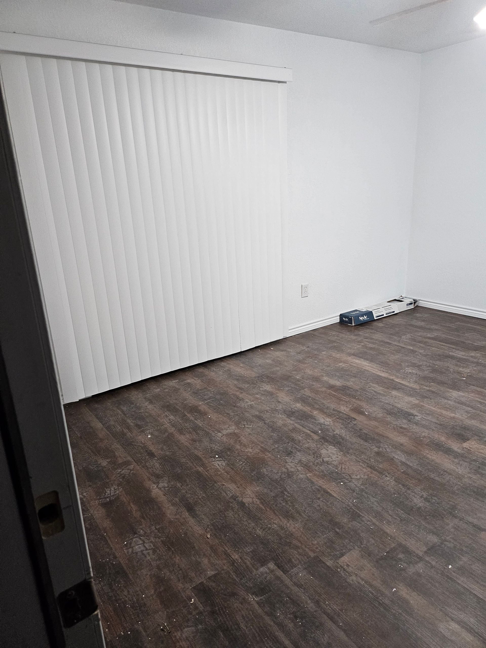 Empty room with wood-look flooring, vertical blinds, and white walls. A few objects are on the floor.