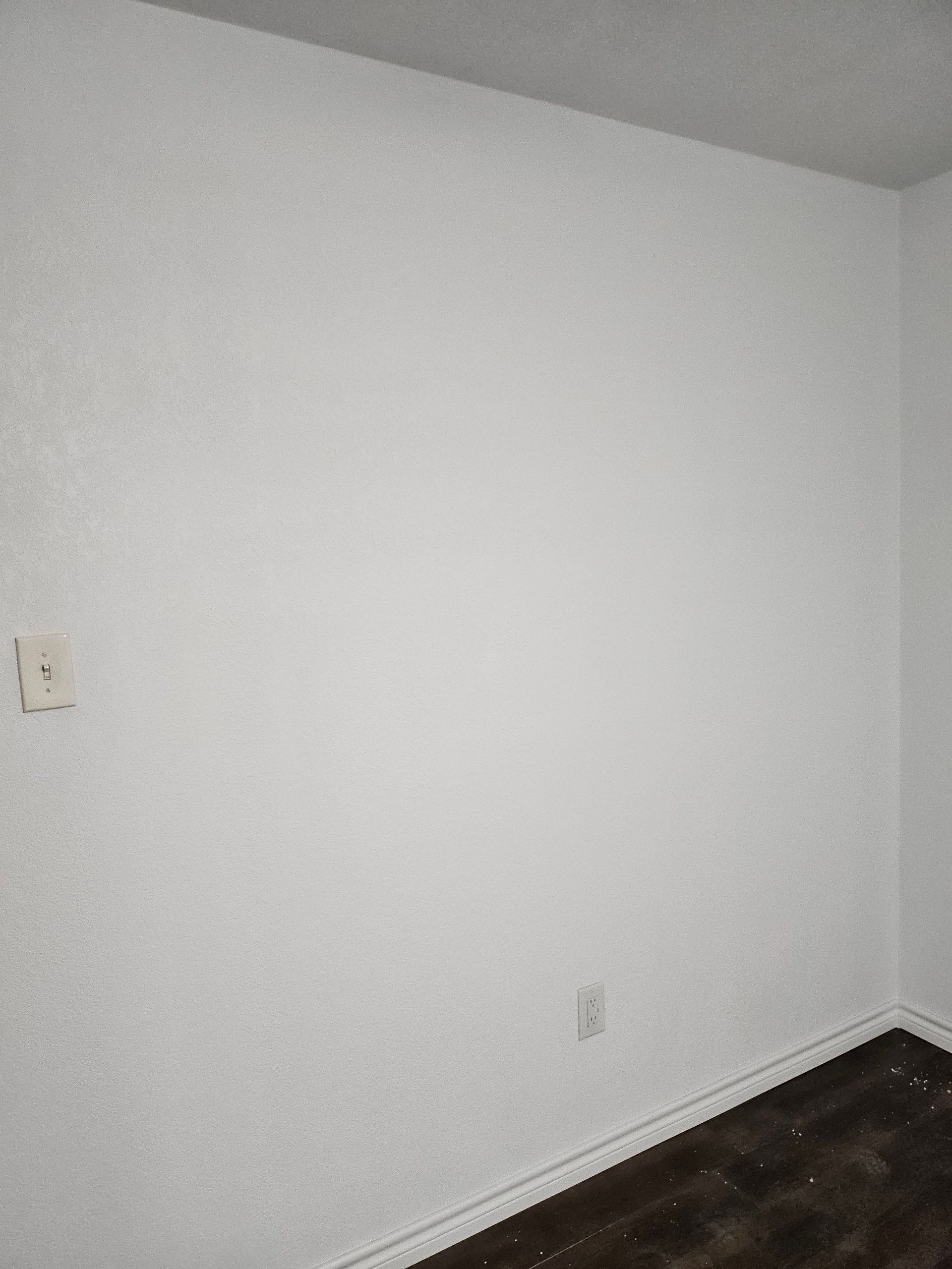 White textured wall in a corner, with a light switch and outlet visible, dark wood-look floor.