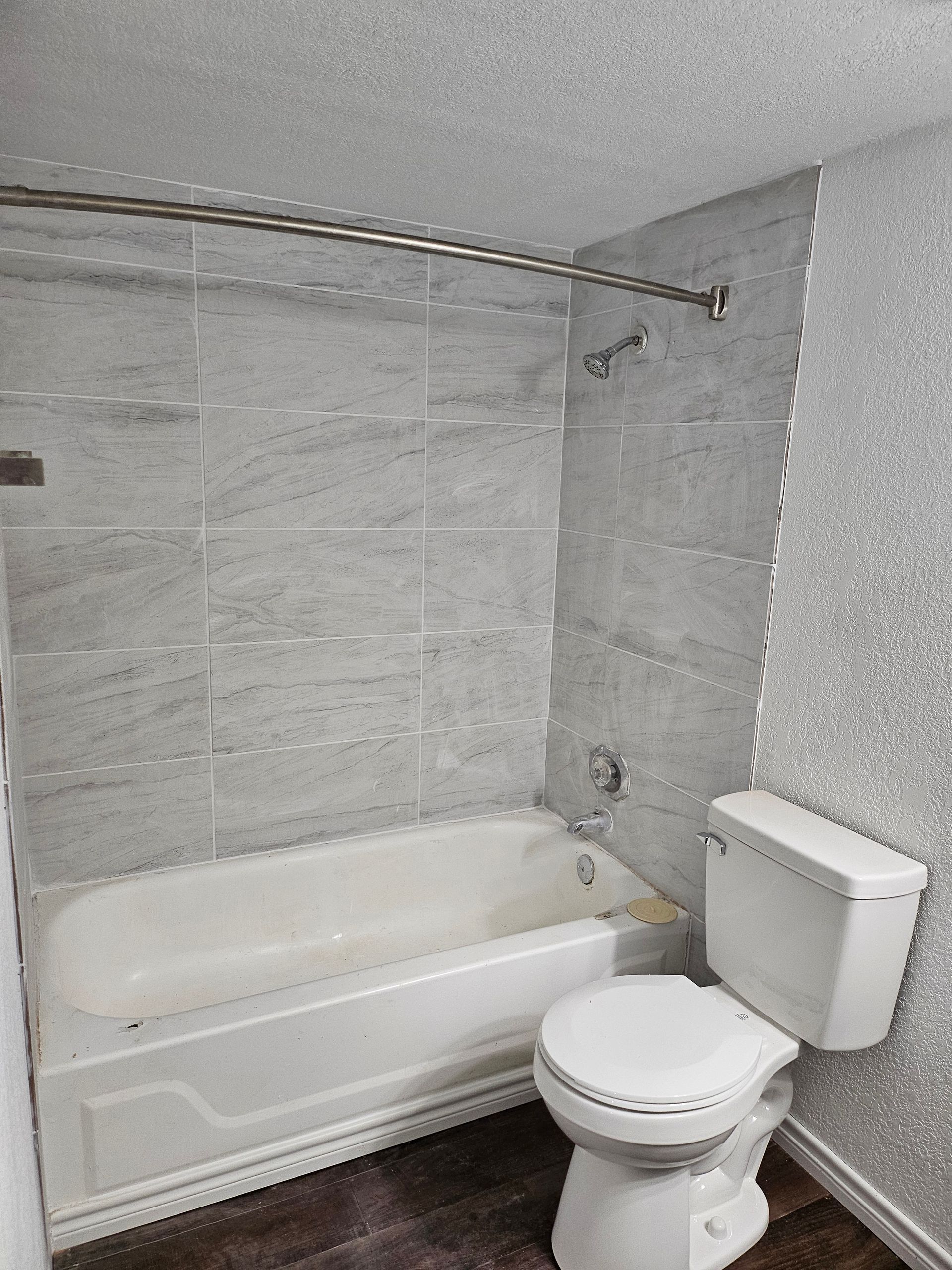 Bathroom with bathtub, toilet, and gray tiled walls. Shower curtain rod is in place.