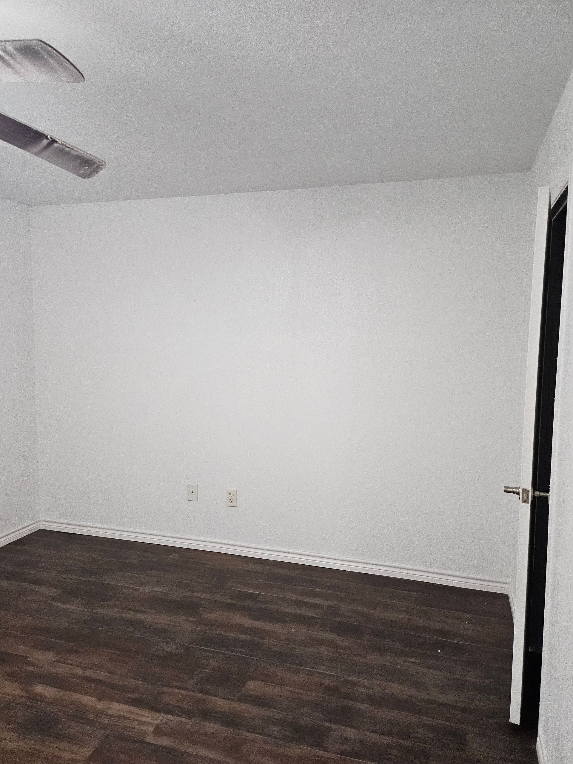 Empty room with white walls, brown floor, ceiling fan, and black door frame.