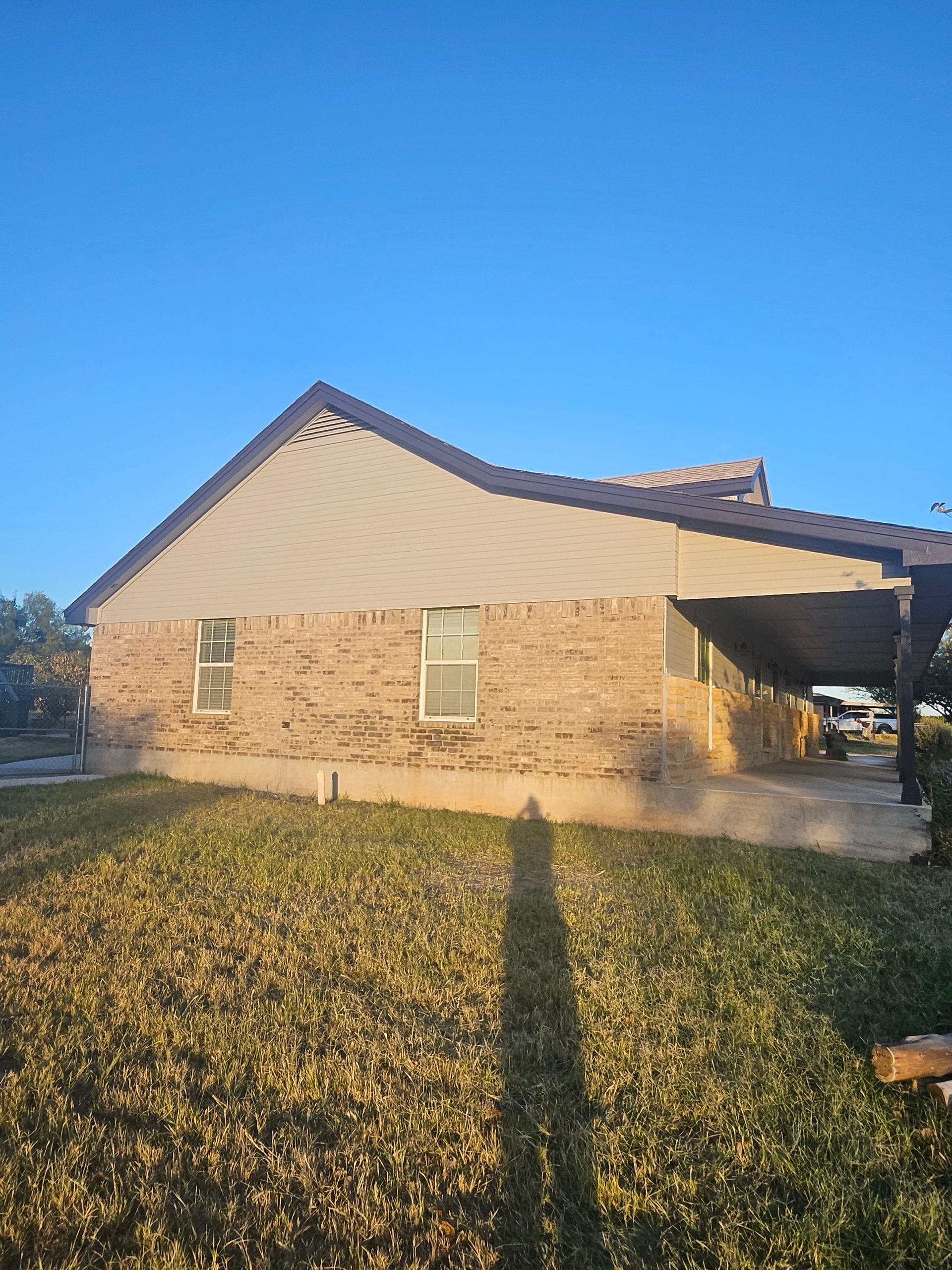 Tan brick house with a dark roof and a covered patio, set against a blue sky.