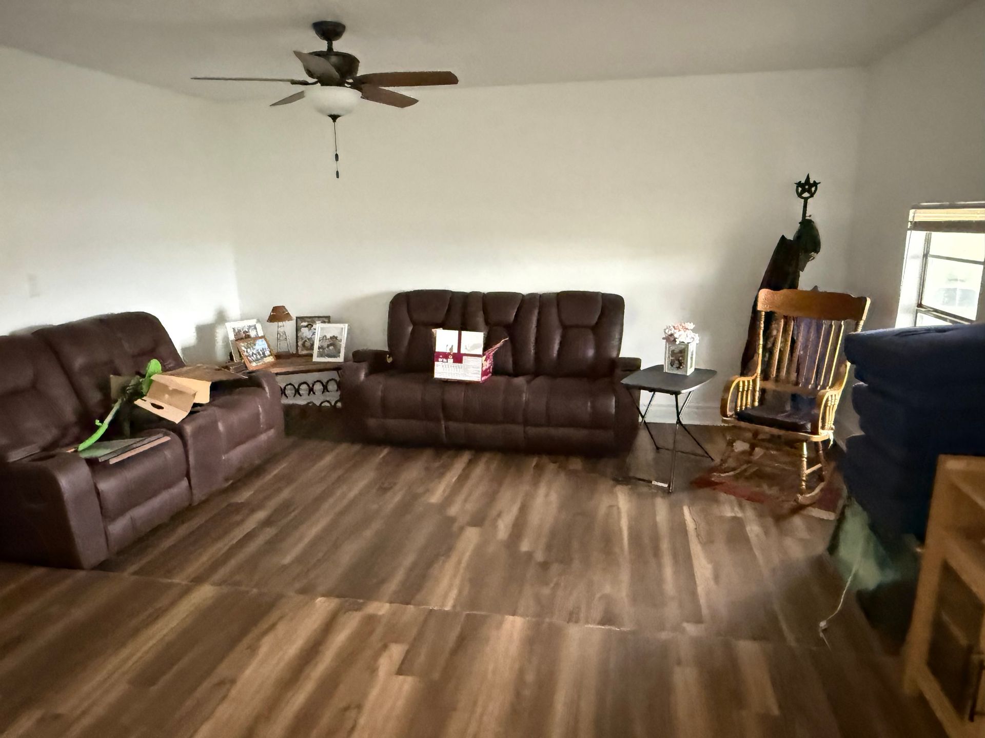 Living room with brown leather furniture, rocking chair, and wood-look flooring. White walls and ceiling fan.