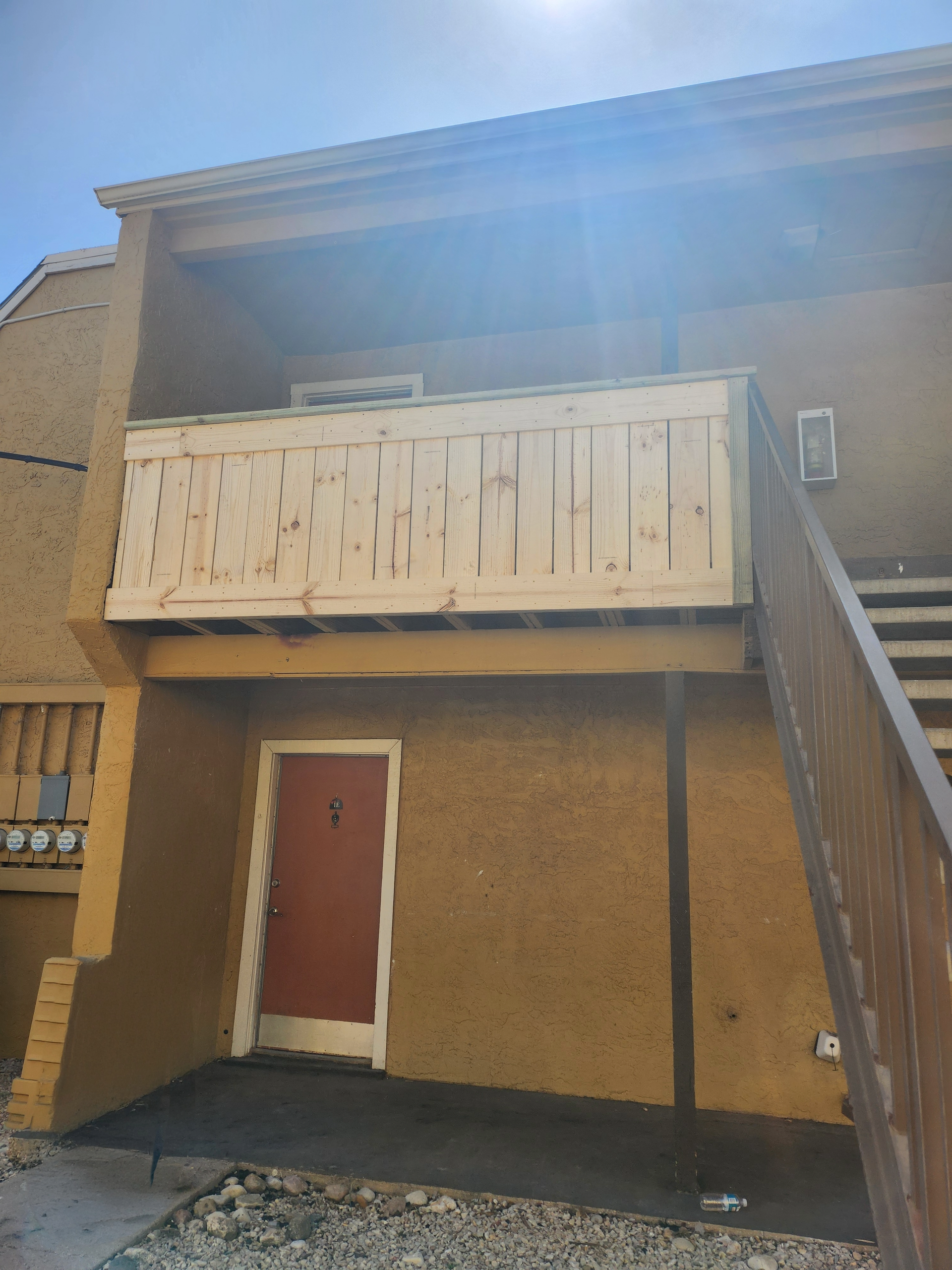 Exterior of a yellow building with a balcony and a red door beneath it. A wooden staircase is visible.