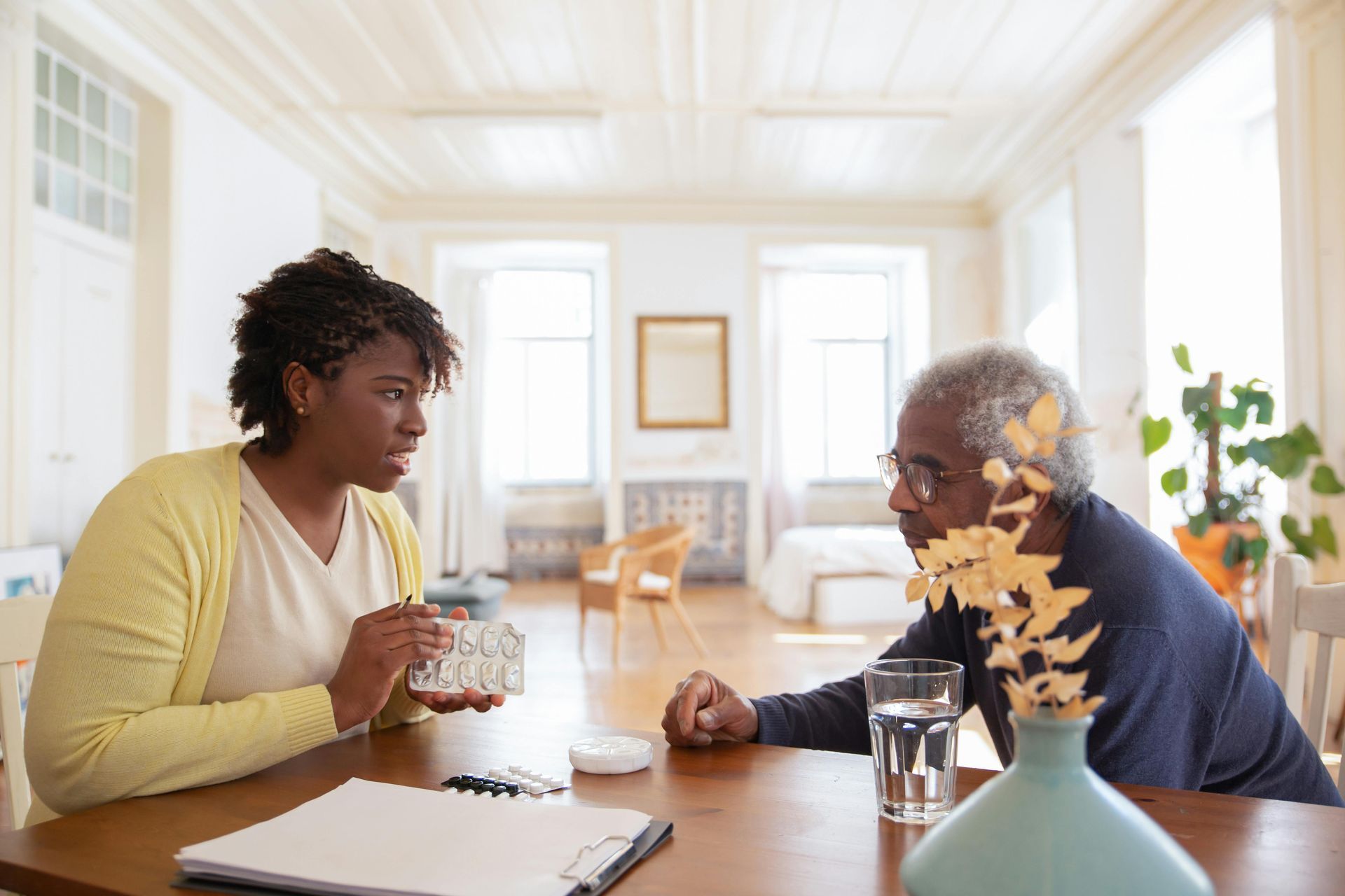 Woman explains medication to a person. They sit at a table in a bright room.