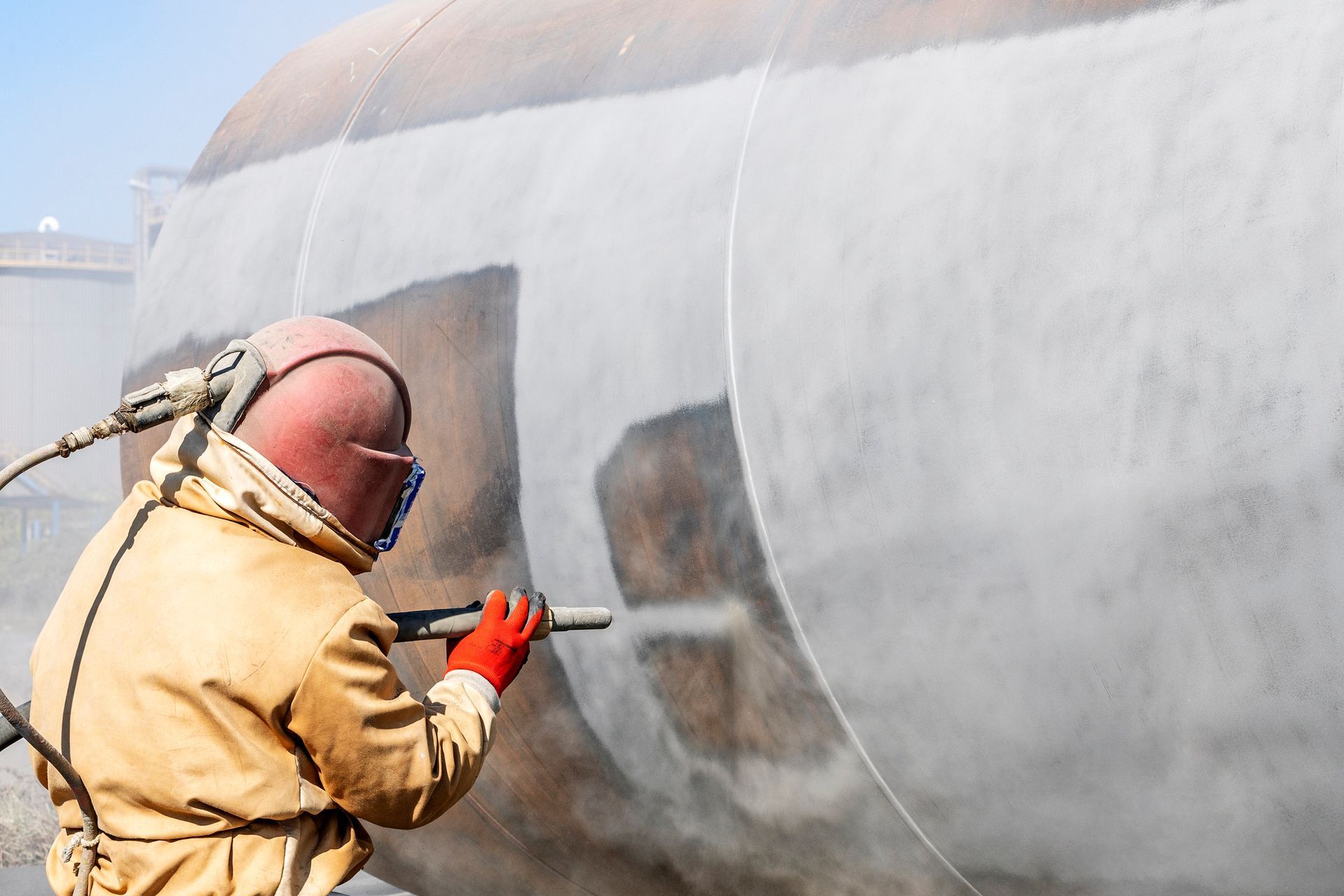A worker wearing a protective, yellow uniform, performs sandblasting service before coating.