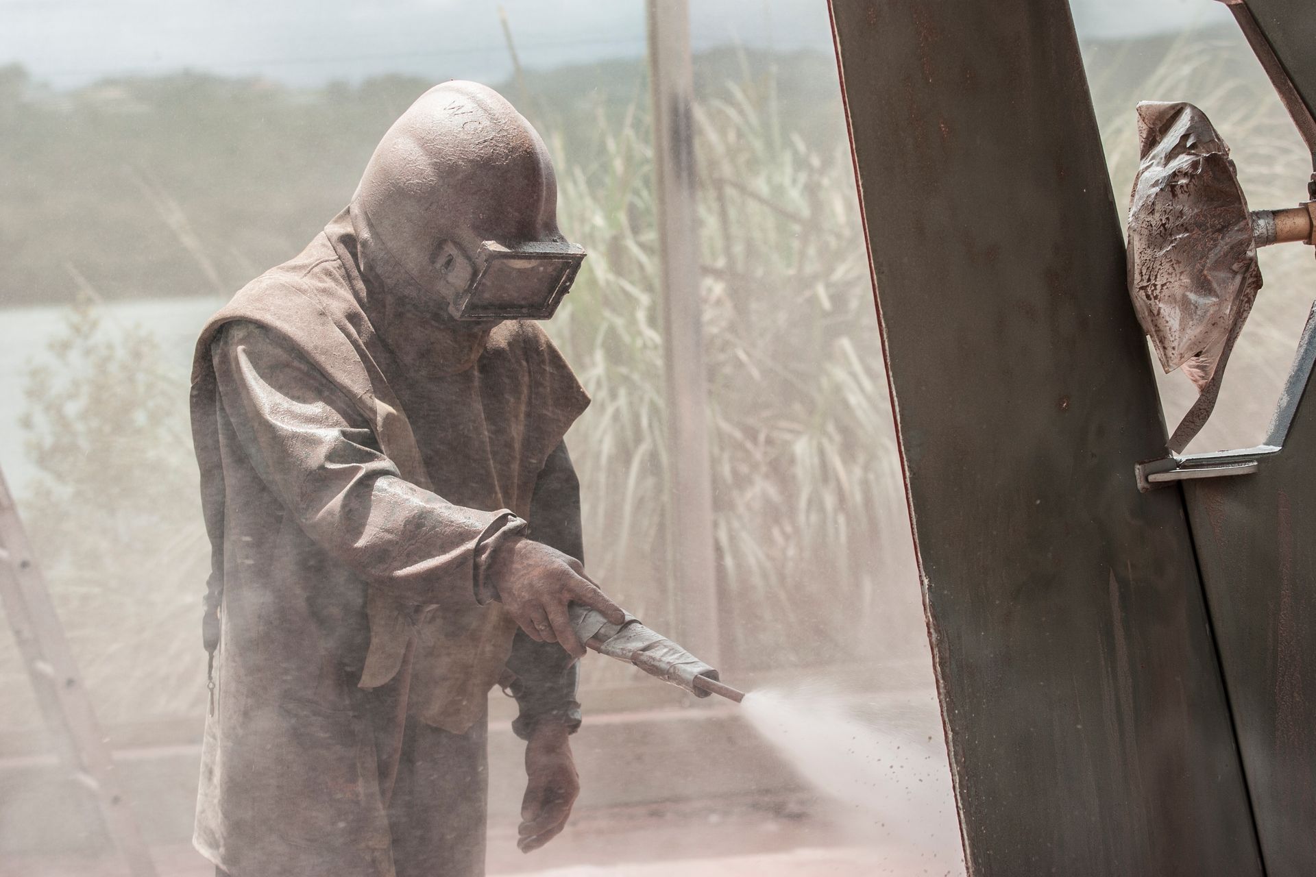 Worker in full gear sandblasts large metal surface outdoors.