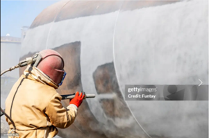 A worker wearing a protective, yellow uniform, performs sandblasting service before coating.