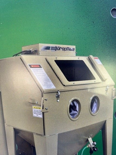 Tan sandblasting cabinet with a black window, two glove holes, and a green background.